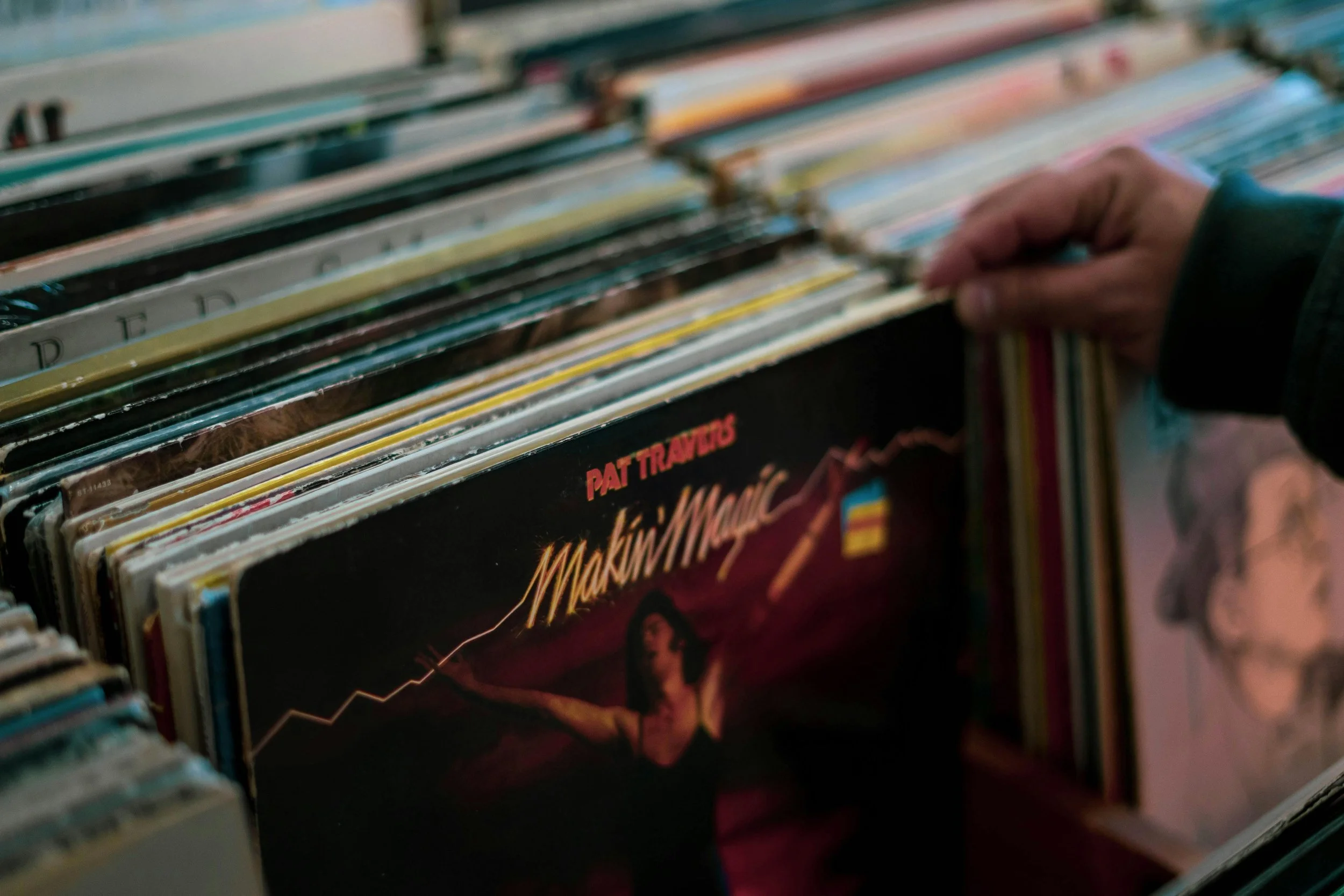 Close-up of a hand reaching into a vinyl record album bin, with various album covers visible.