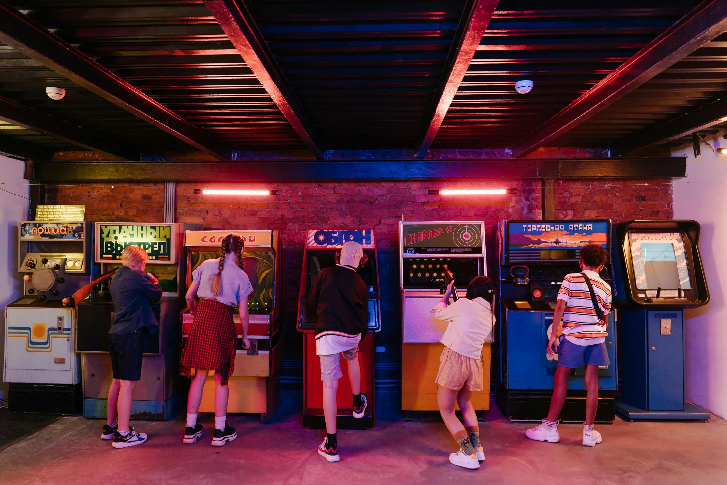 Six children playing arcade games against a brick wall in a dimly lit arcade. Neon lights cast a colorful glow.