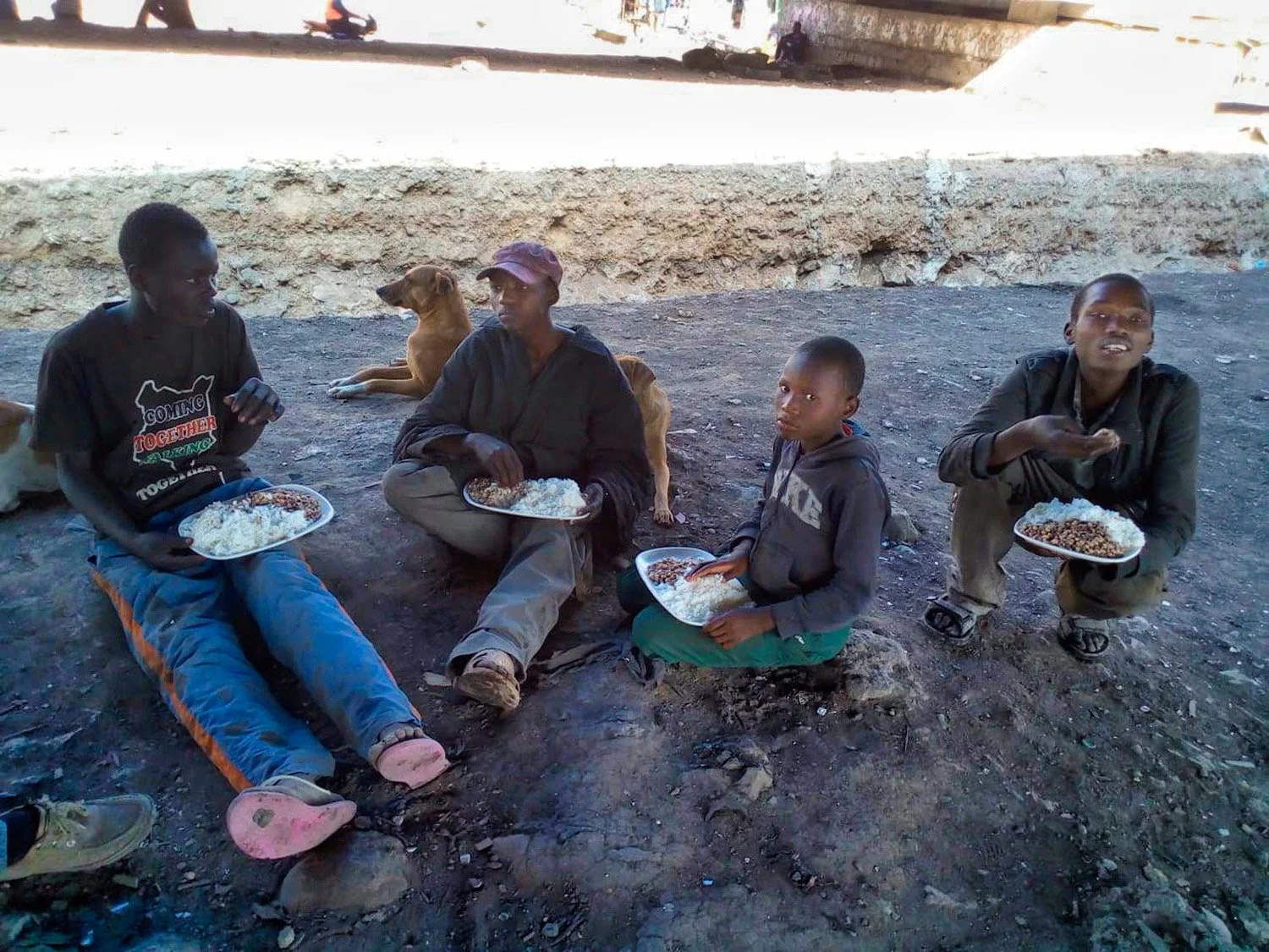 Vier Jugendliche sitzen mit zwei Hunden zusammen am Boden unter einer Brücke und essen aus Blechtellern. Die Nahrung wurde ihnen von der Hilfsorganisation Nafrika gespendet