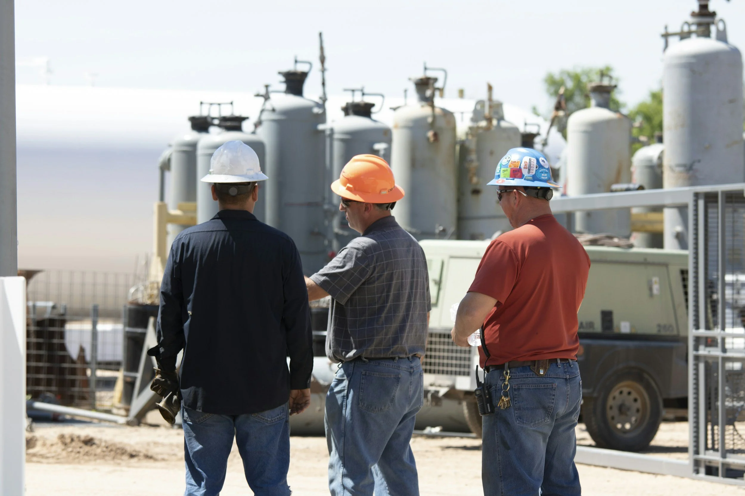 Three field workers in safety helmets standing beside industrial equipment during an on-site discussion.