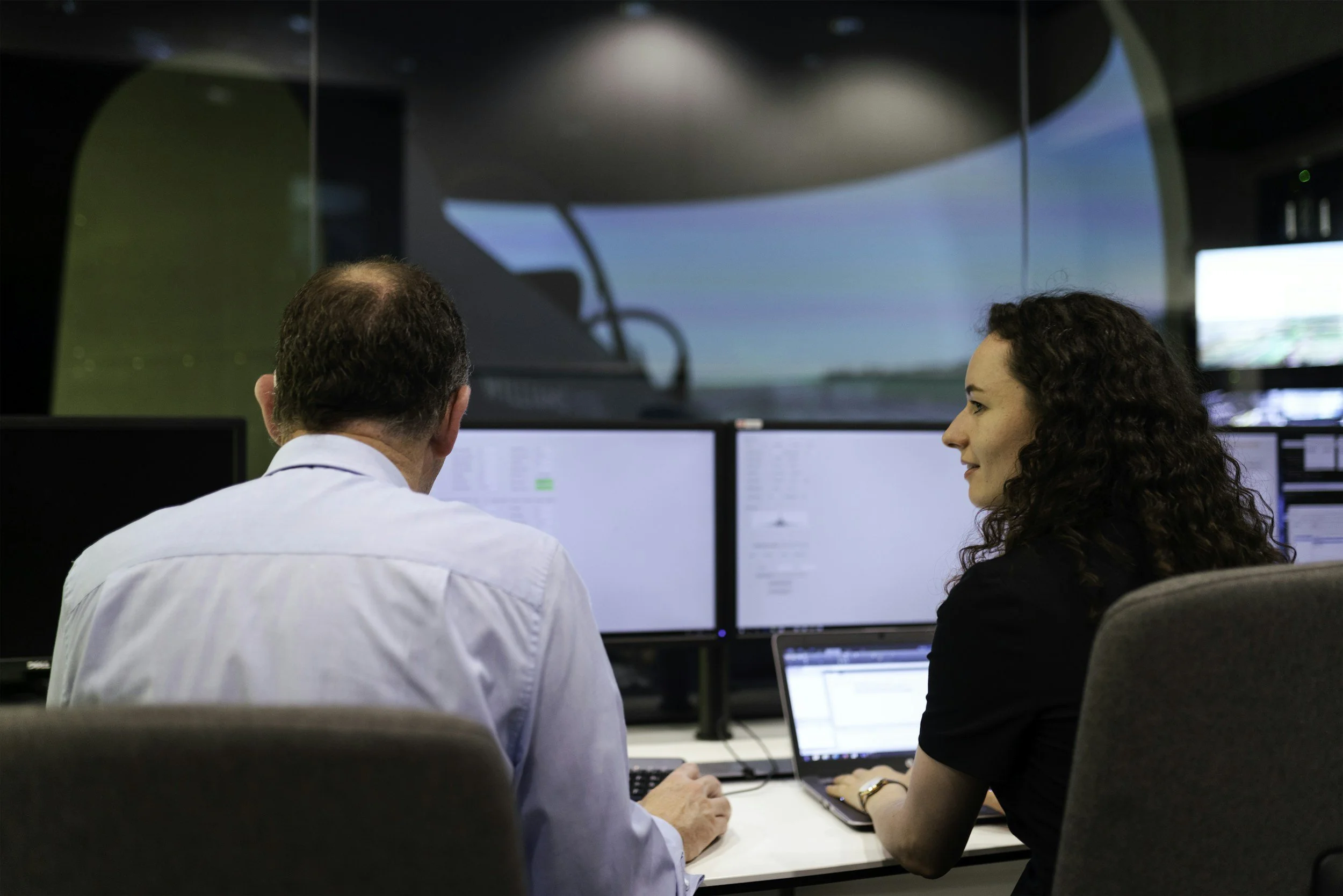 Two people in a control room reviewing industrial system data across multiple monitors.