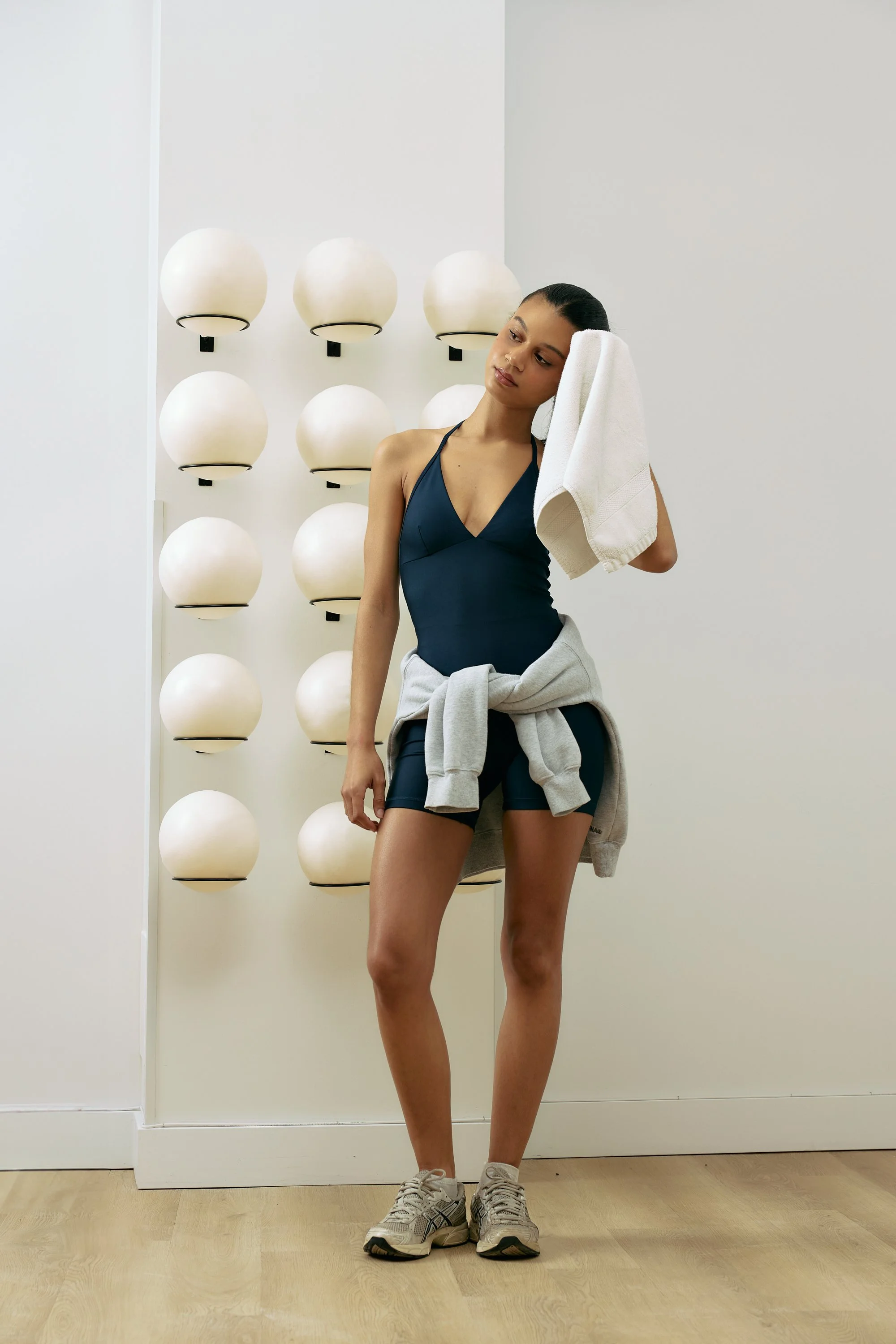 A young woman in workout attire, holding a towel to her head, standing in a minimalistic room with spherical wall lights.