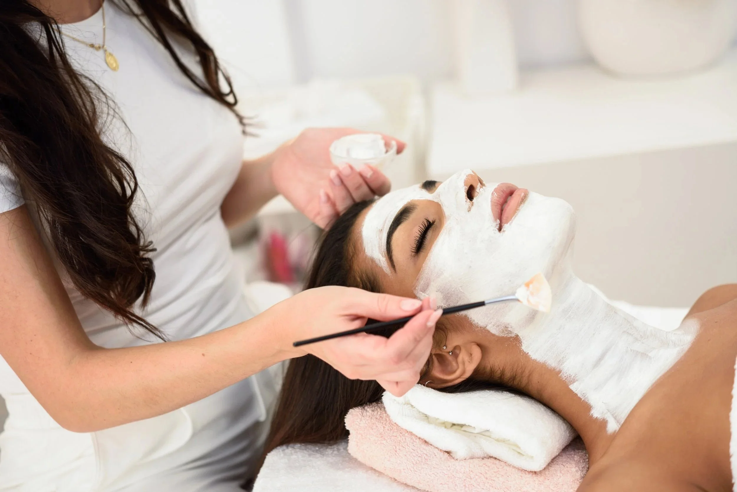 Woman receiving a facial treatment at a spa, with white facial mask applied and lying on a towel