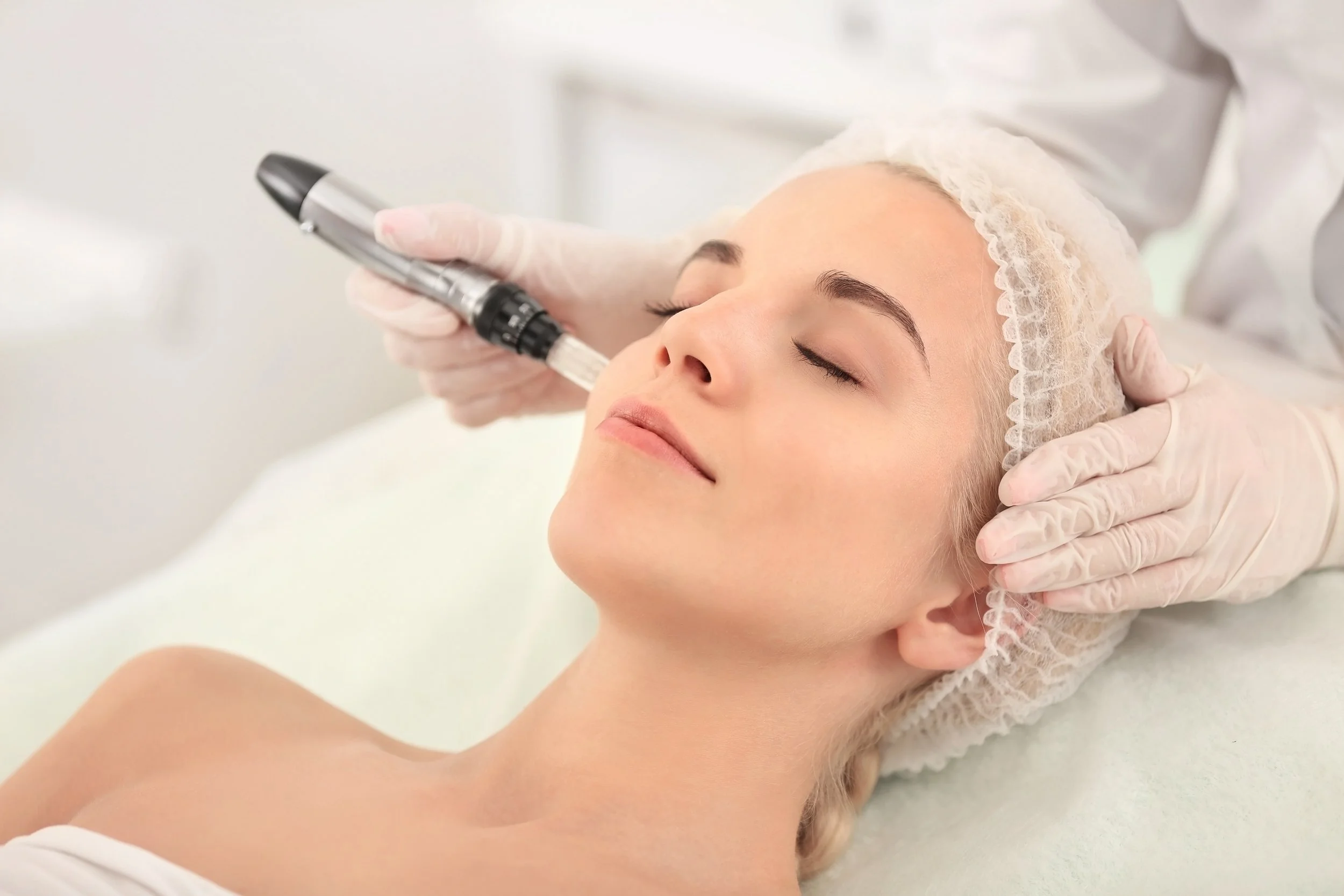 A woman is receiving a facial treatment with a microdermabrasion device in a skincare clinic, lying covered with a hair cap and with her eyes closed.