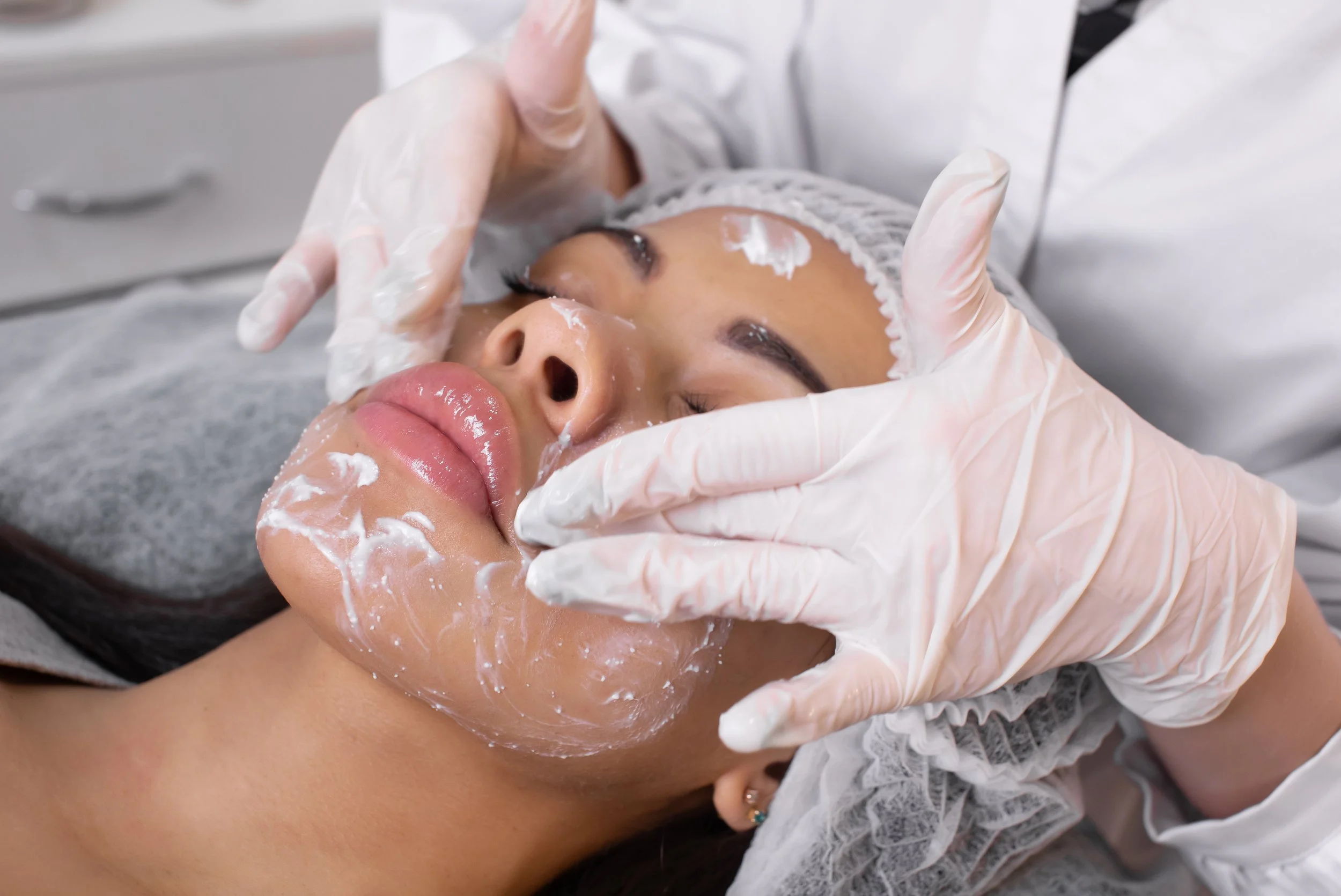 A woman receiving a facial treatment from a professional wearing gloves, with facial cream applied and a headband on.