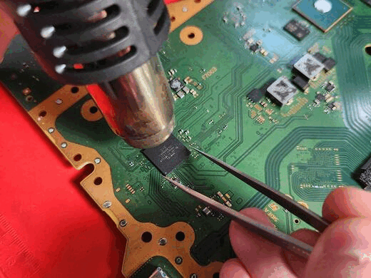 A person uses a soldering iron and tweezers to repair a green printed circuit board with various electronic components.