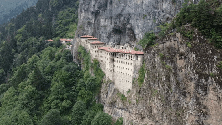 A monastery built into a cliffside with multiple levels and red-tiled roofs, surrounded by dense green forest and rock formations.