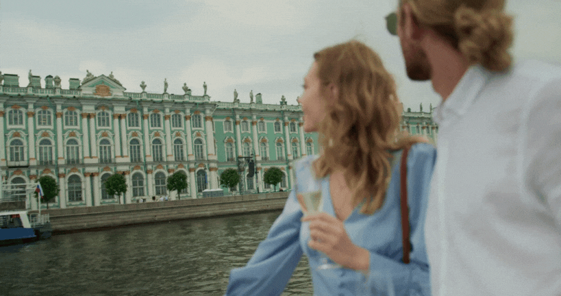 A man and a woman standing by a river with an ornate, pastel-colored building in the background. The woman is holding a glass of wine and looking at the building.