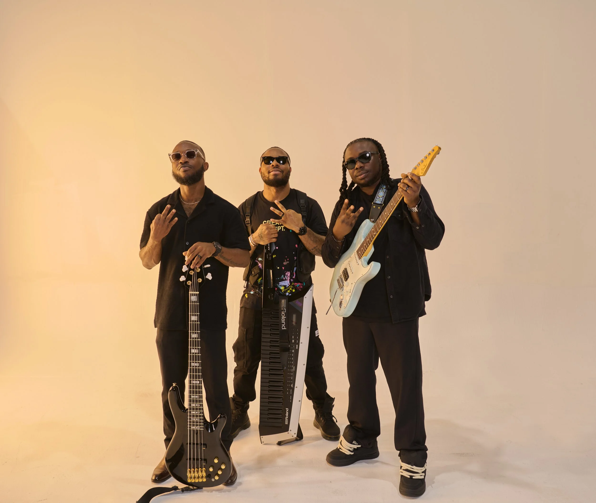 Three men posing with musical instruments on a plain background: one with a bass guitar, one with a keyboard, and one playing an electric guitar.