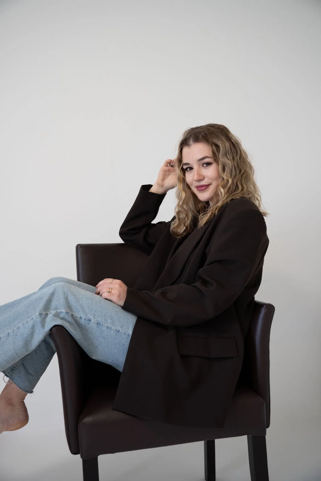 Junge Frau sitzt lässig auf einem Stuhl, trägt schwarze Jacke und Jeans, lächelt und schaut in die Kamera, vor hellem Hintergrund.