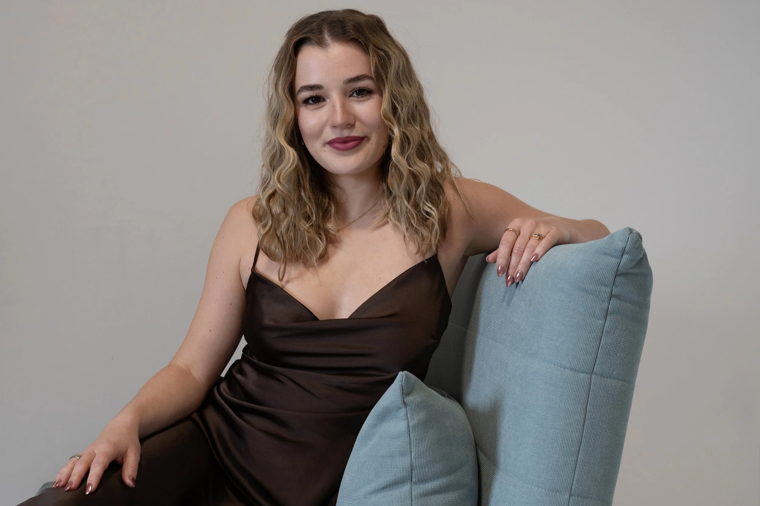 Junge Frau mit Locken sitzt auf einem blauen Sofa vor einem grauen Hintergrund, trägt ein braunes Kleid, trägt Ringe, eine Halskette, und sieht freundlich in die Kamera.
