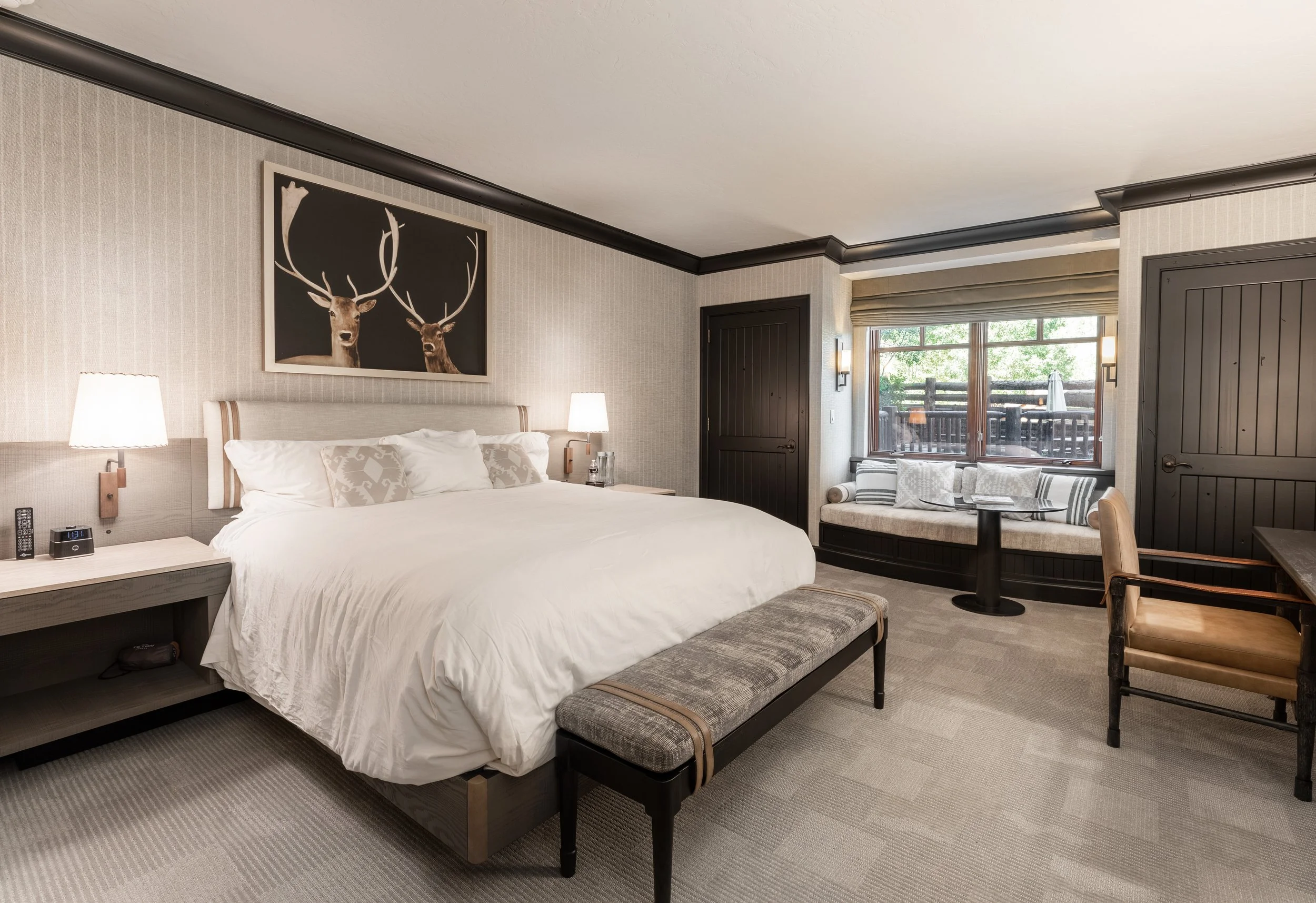 A Ritz-Carlton Suite with a large bed, two lamps, a window seat with pillows, and a painting of two deer with antlers on the wall.