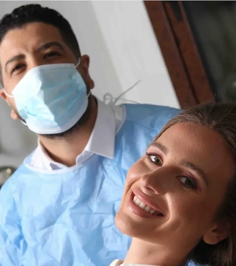 A smiling woman with light brown hair lying back on a medical chair, and a man wearing a blue medical gown, face mask, and white shirt standing behind her.