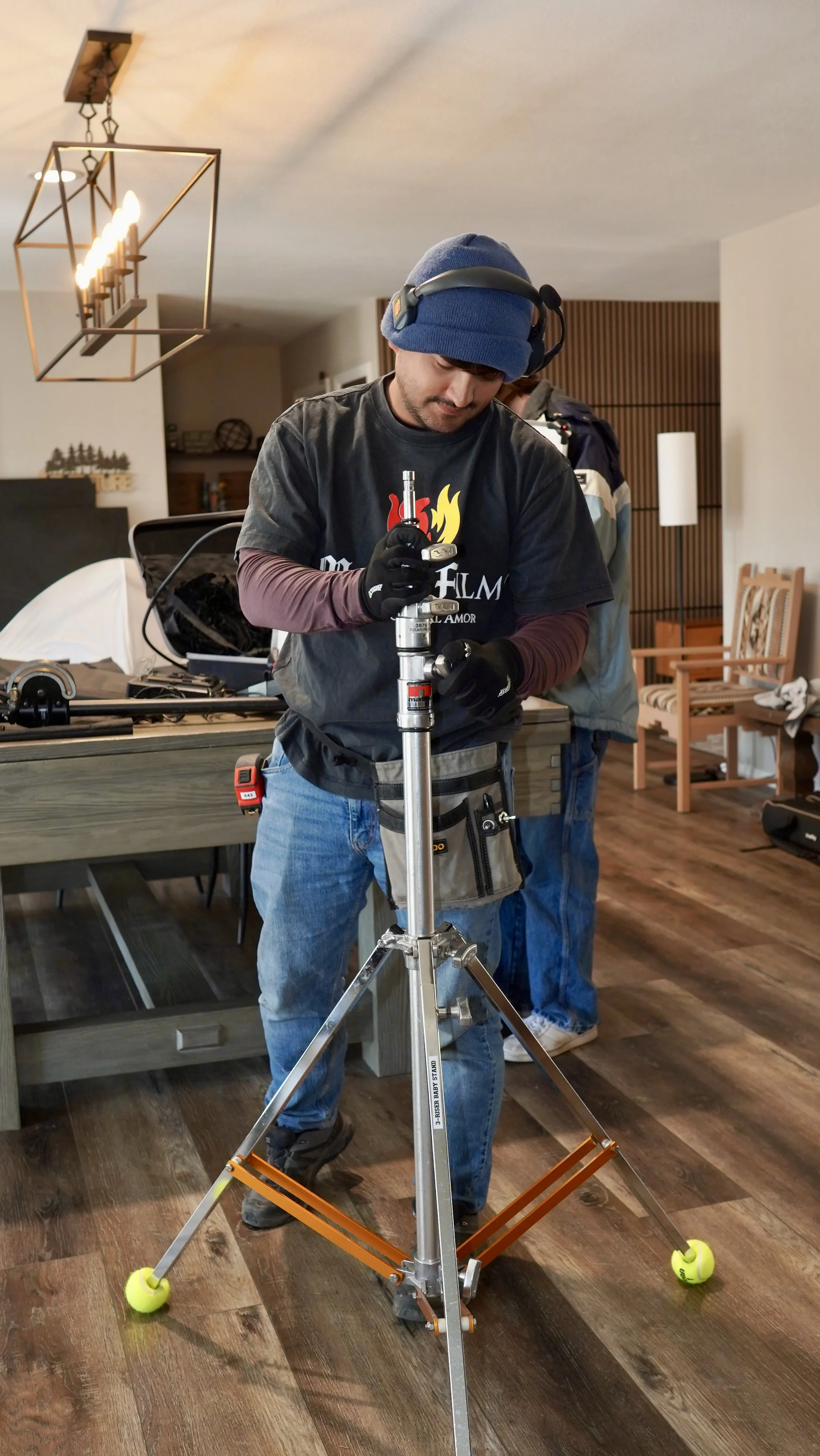 A man wearing a blue beanie, black gloves, and a black T-shirt working with a camera stabilization rig indoors, with another person in the background.