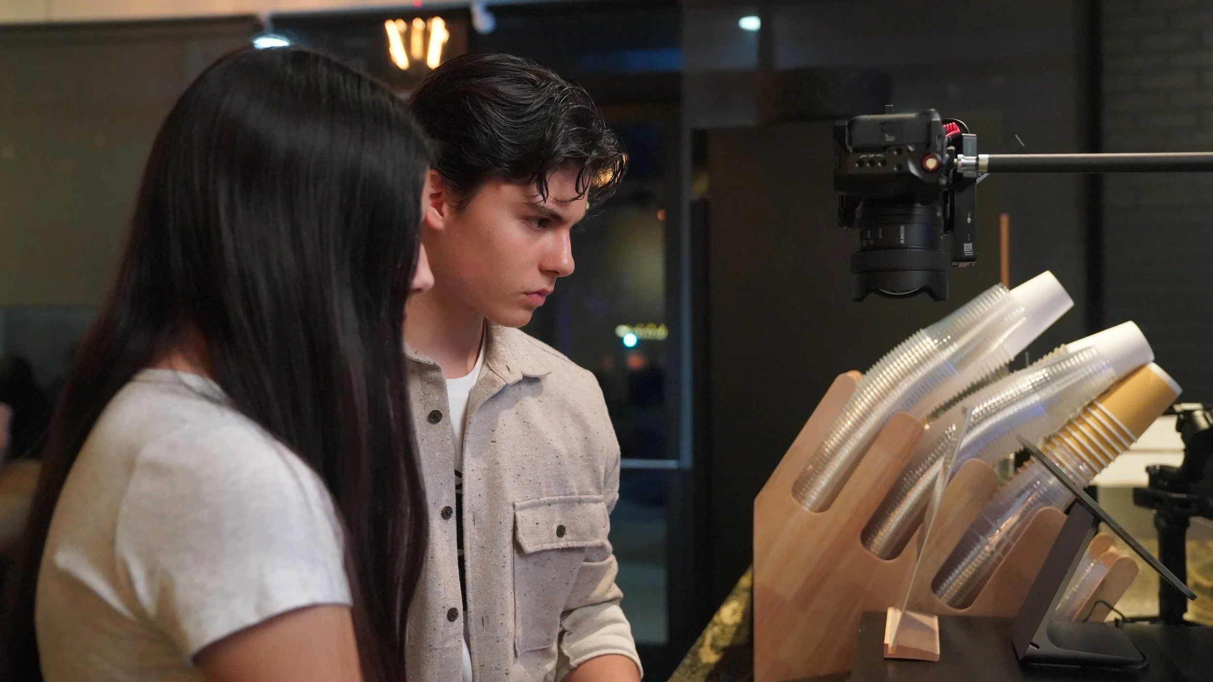 Two people, a woman with long dark hair and a man with short dark hair, looking at a display of plastic cups and a camera on a boom arm in an indoor setting.