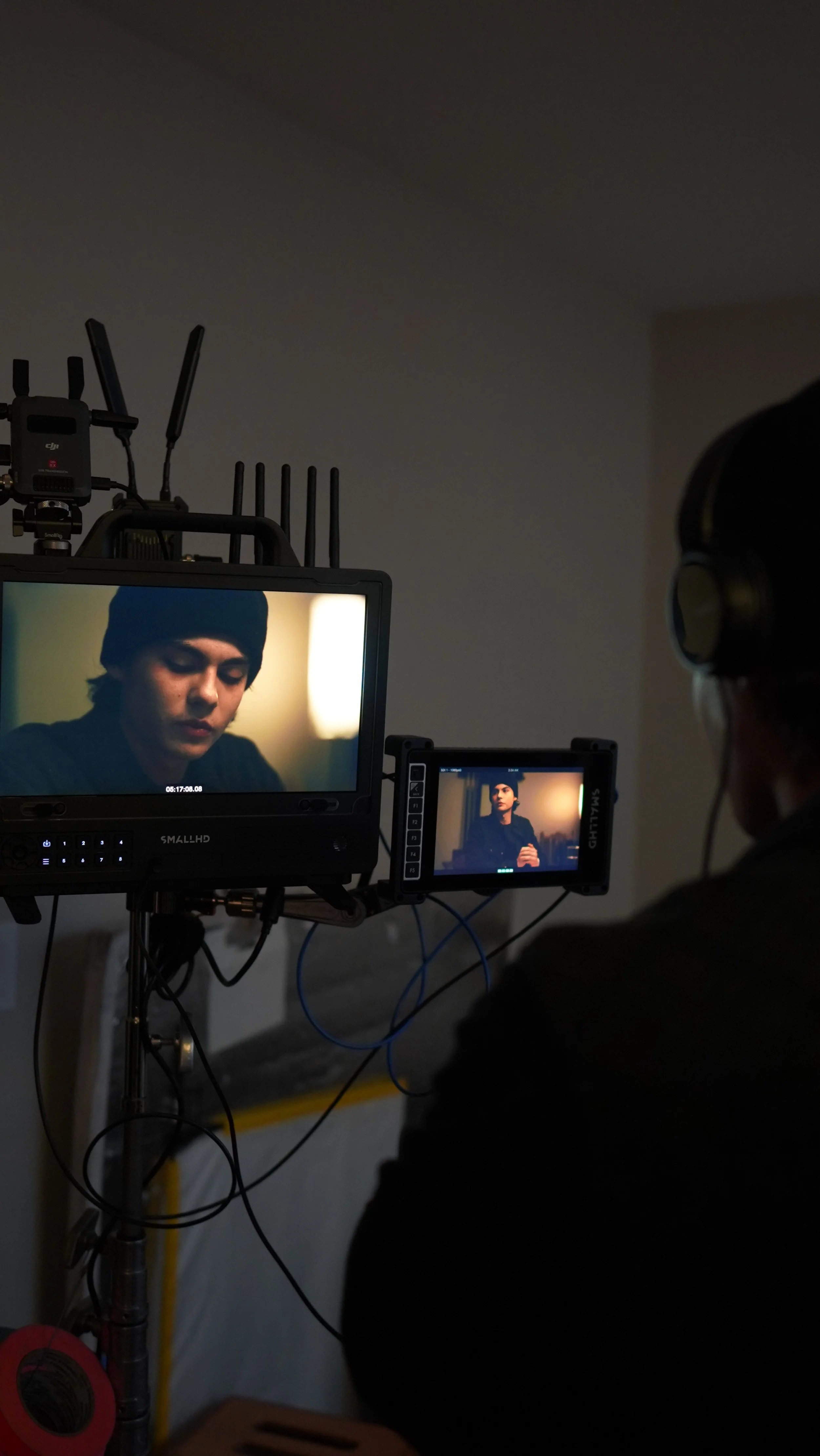 A film director or crew member on set uses monitors to review scenes being shot, with a woman actor visible on the screens, wearing a dark beanie and acting.