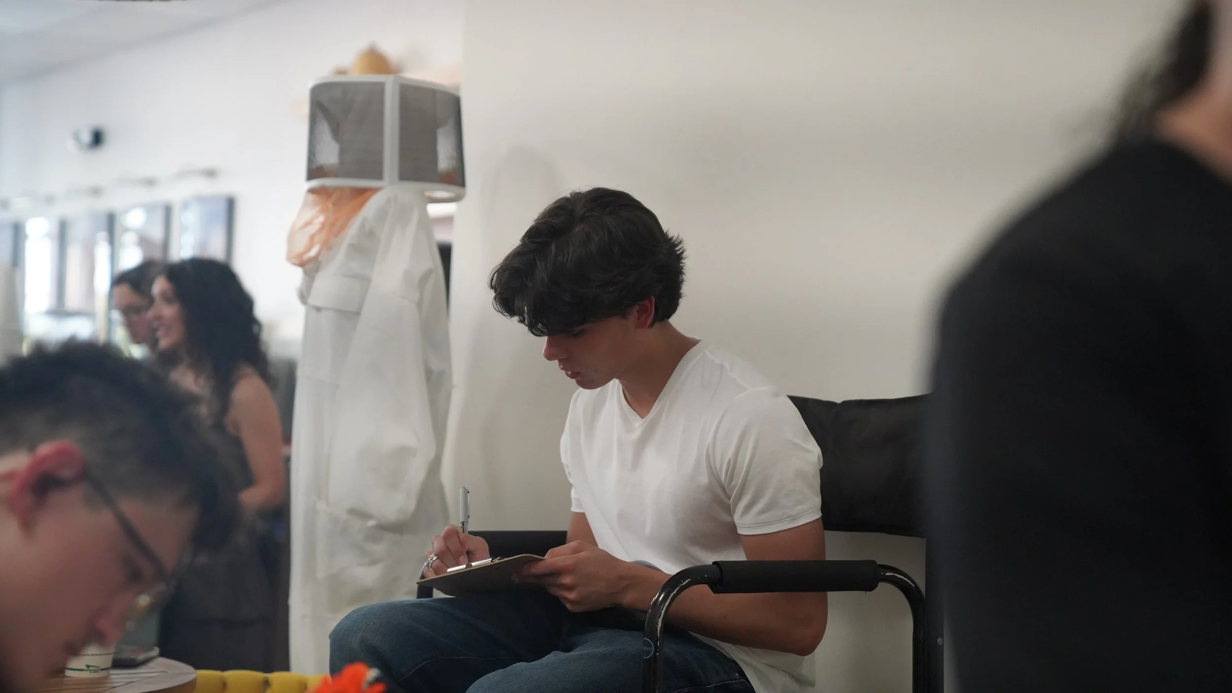 Young man with dark, wavy hair seated on a black chair, writing on a clipboard in a room with other people. In the background, two women are smiling and talking, and a third person is partially visible on the right.