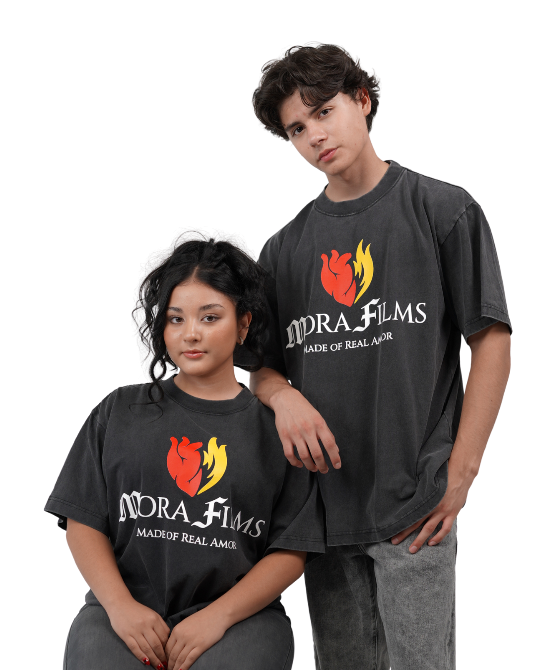A young man and woman wearing matching black T-shirts with a red and yellow heart graphic and the words 'MORA FILMS MADE OF REAL AMOR' printed on them, posing against a black background.