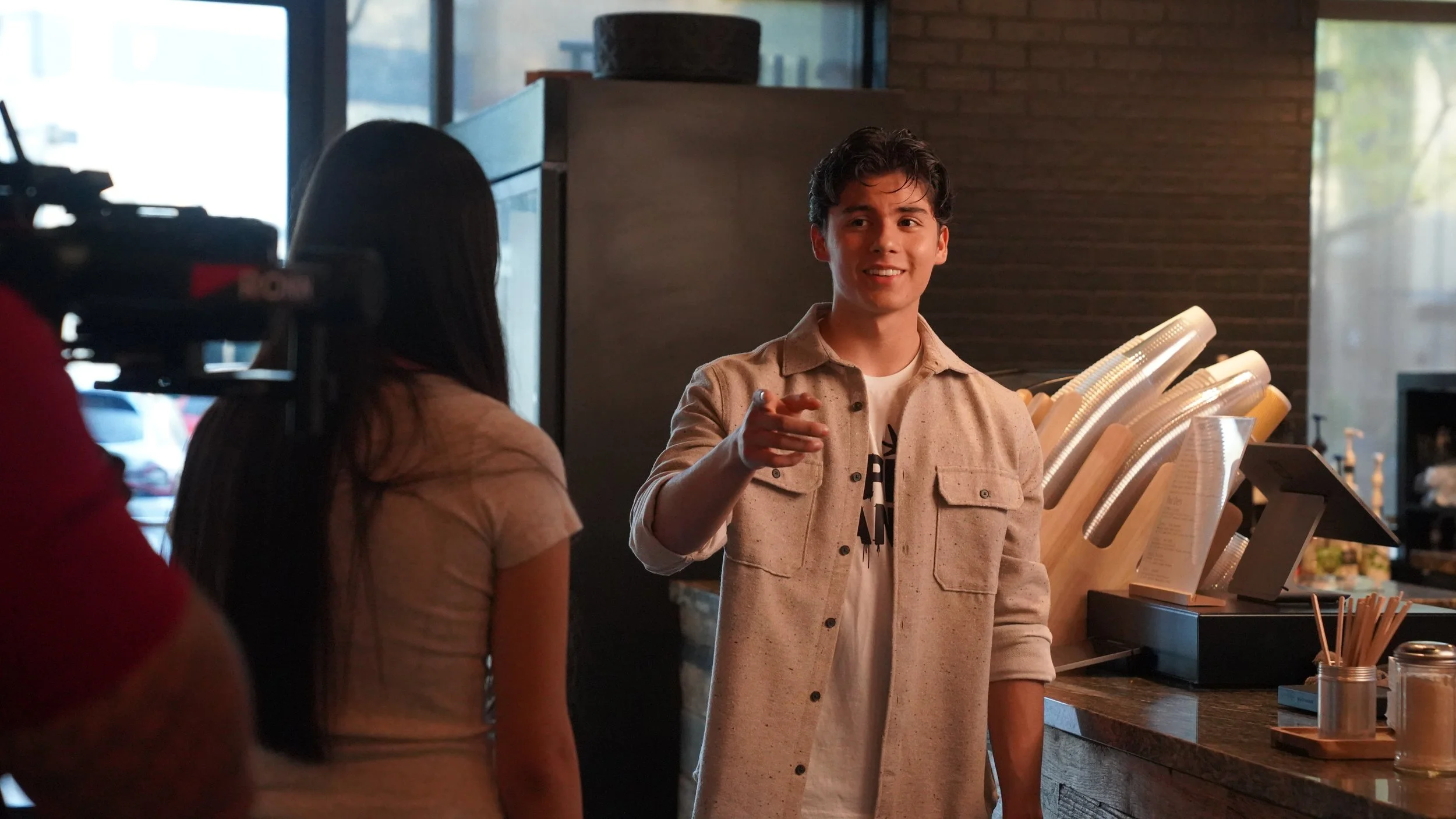 Young man smiling and pointing in a casual cafe or restaurant setting, with two women in front of him and a camera filming him.