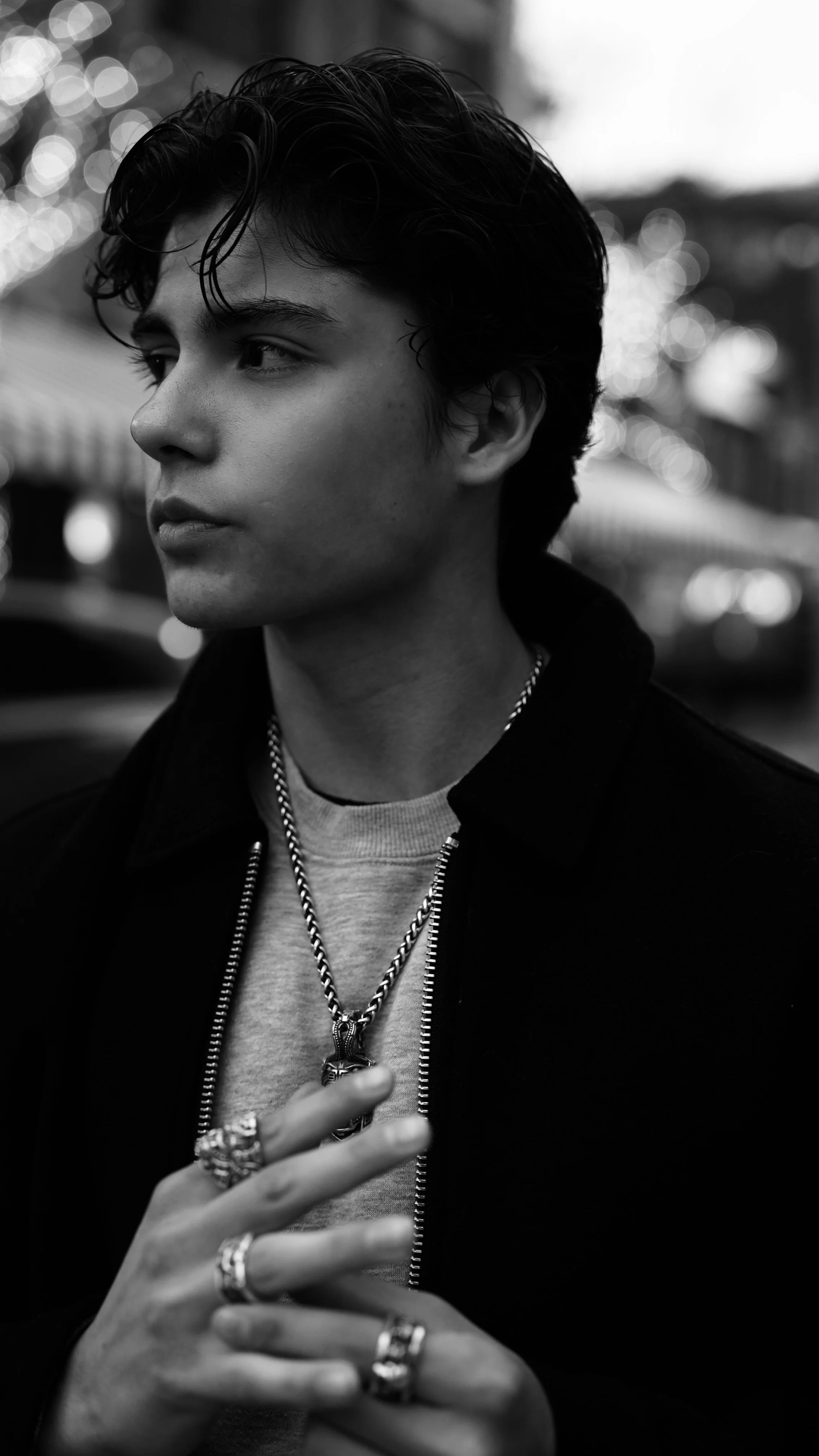 A young man with wavy dark hair, wearing rings, a chain necklace, and a jacket, is looking to the side in an outdoor setting with blurred lights in the background.