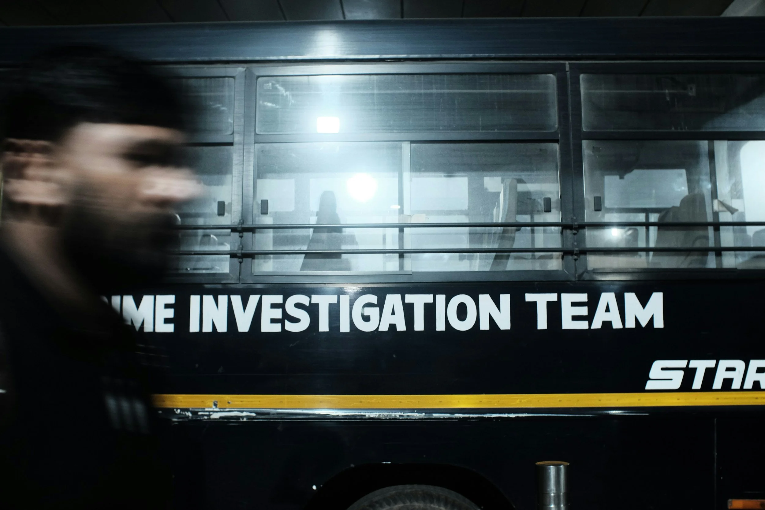 A blurred person walking past a black vehicle labeled 'HOME INVESTIGATION TEAM' with a yellow stripe, parked indoors.