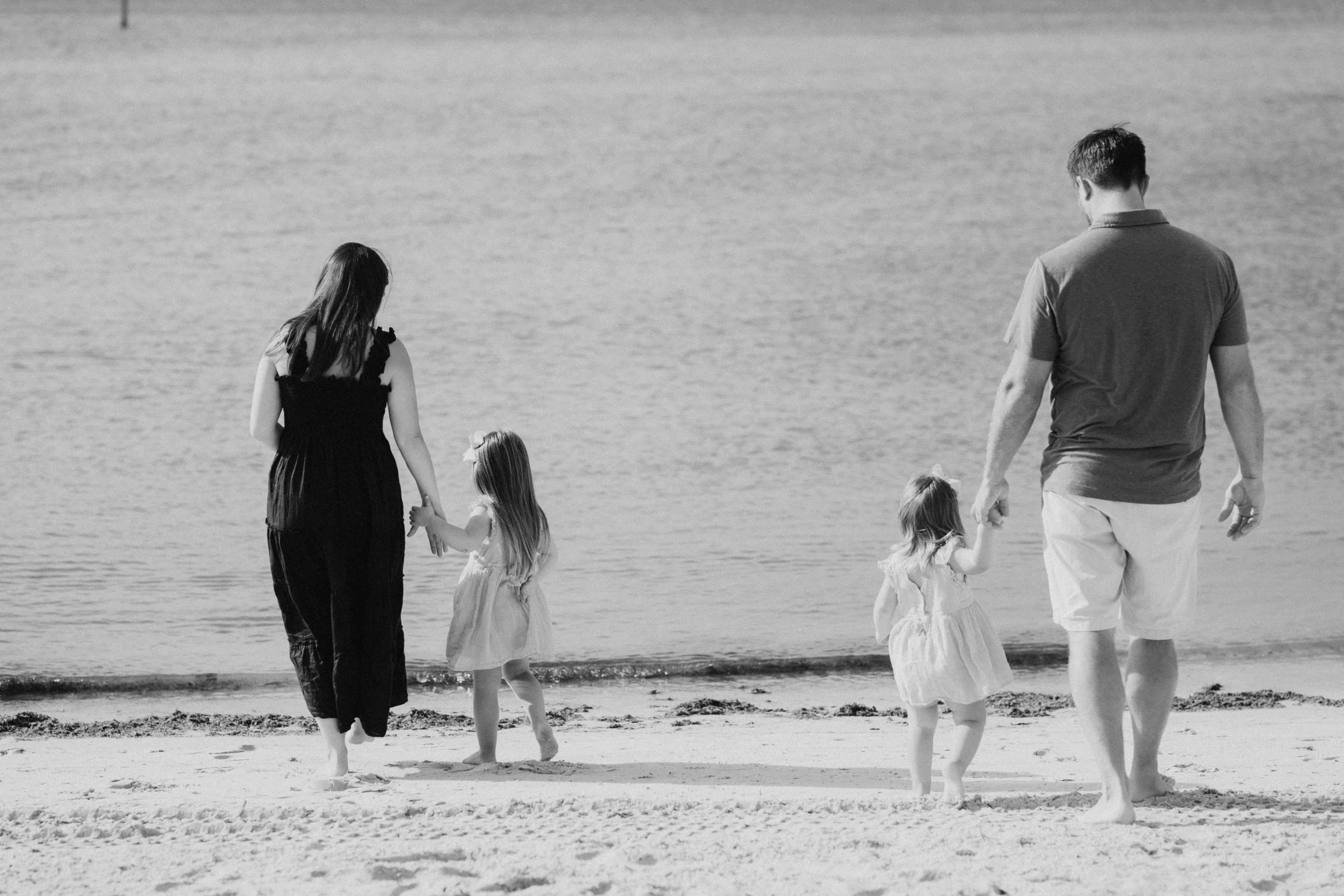 A family with two adults and two young girls walking on the beach near the water, holding hands.