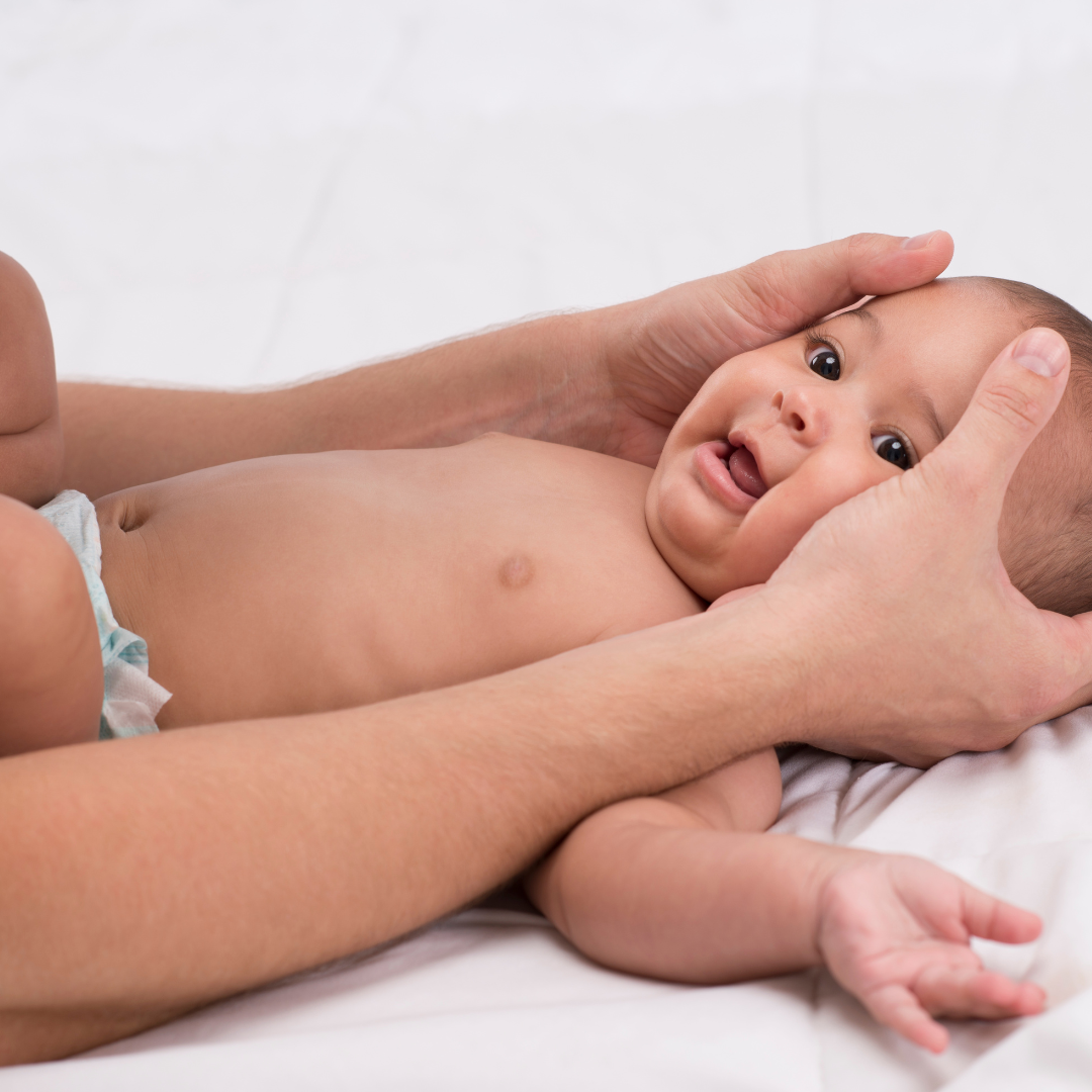 A baby lying on a bed, being gently held and supported by an adult's hands, with the baby looking up with a curious expression.