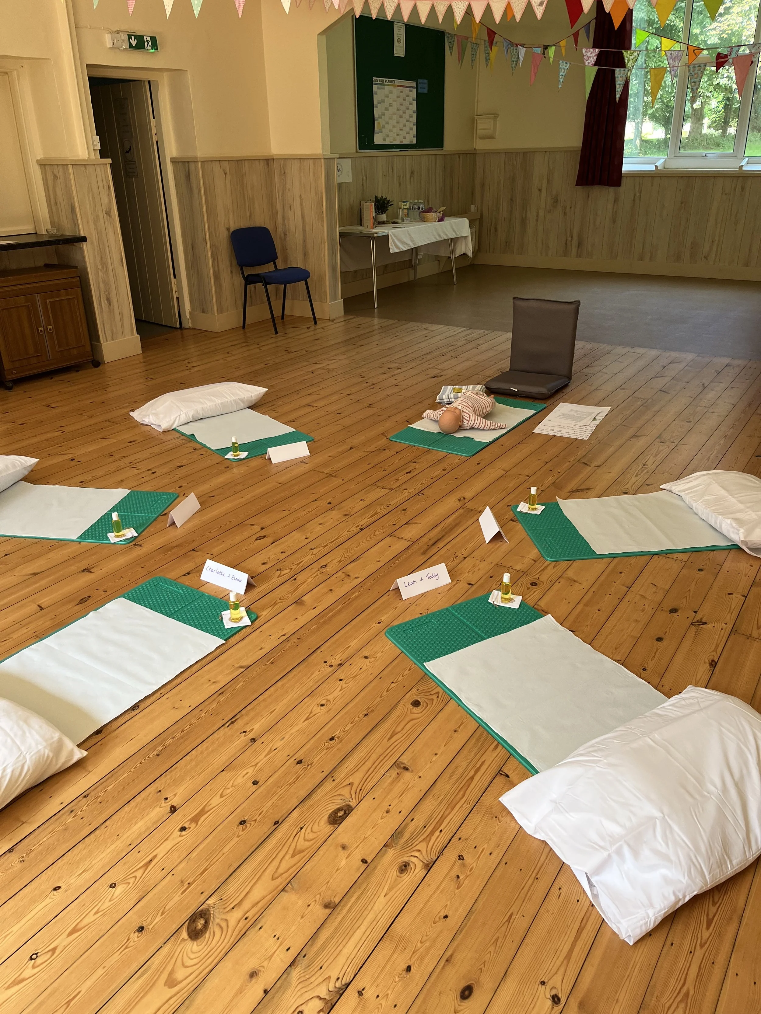 A room set up with five mats, each with a pillow,  arranged in a semi-circle. In the center, there is a doll on a mat with a trainer's seat nearby. The room has wooden floors, decorated with colorful paper bunting hanging from the ceiling, and a table with refreshments in the background.