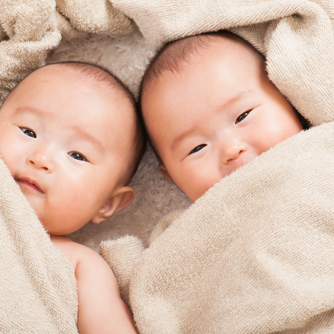Two babies wrapped in beige towels lying side by side on a soft, textured blanket.
