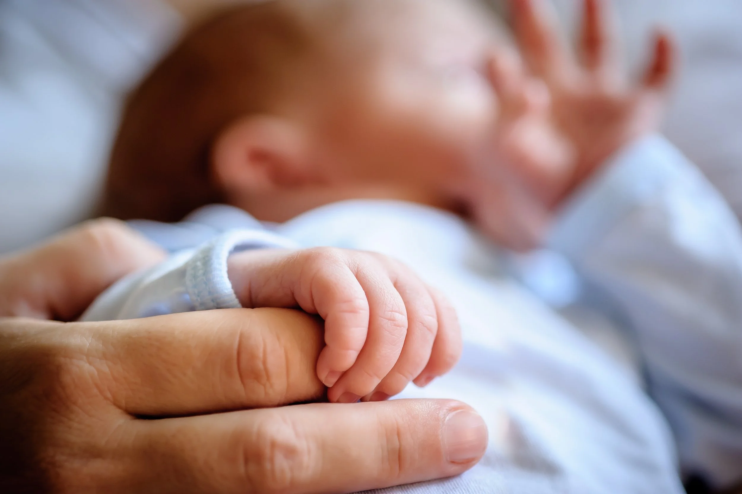 Close-up of a newborn baby's hand grasping an adult's index finger, with the baby's face blurred in the background.