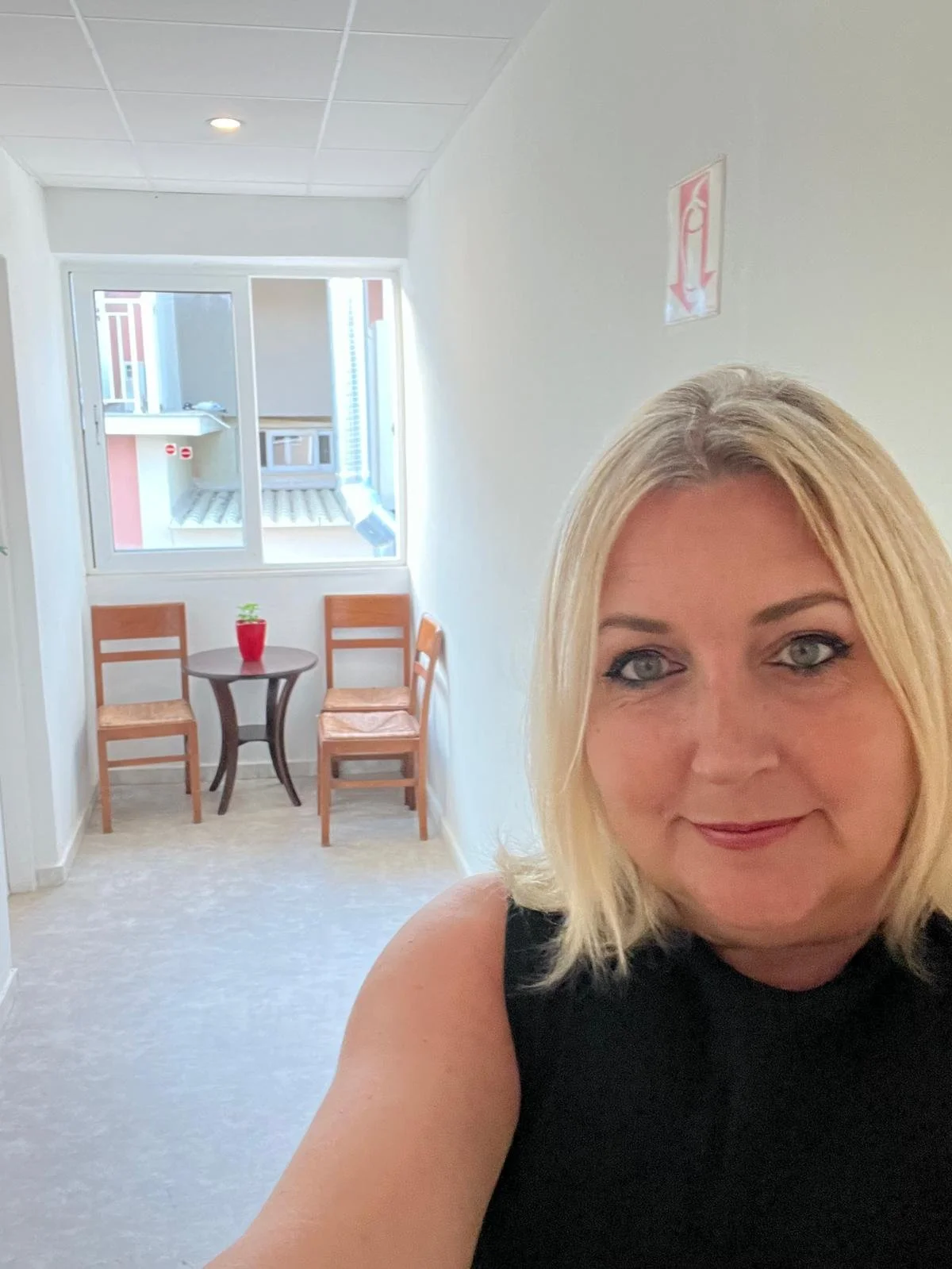 A woman with blonde hair wearing a black top taking a selfie in a bright hallway with a small table, two chairs, and a window with a view of neighboring buildings.