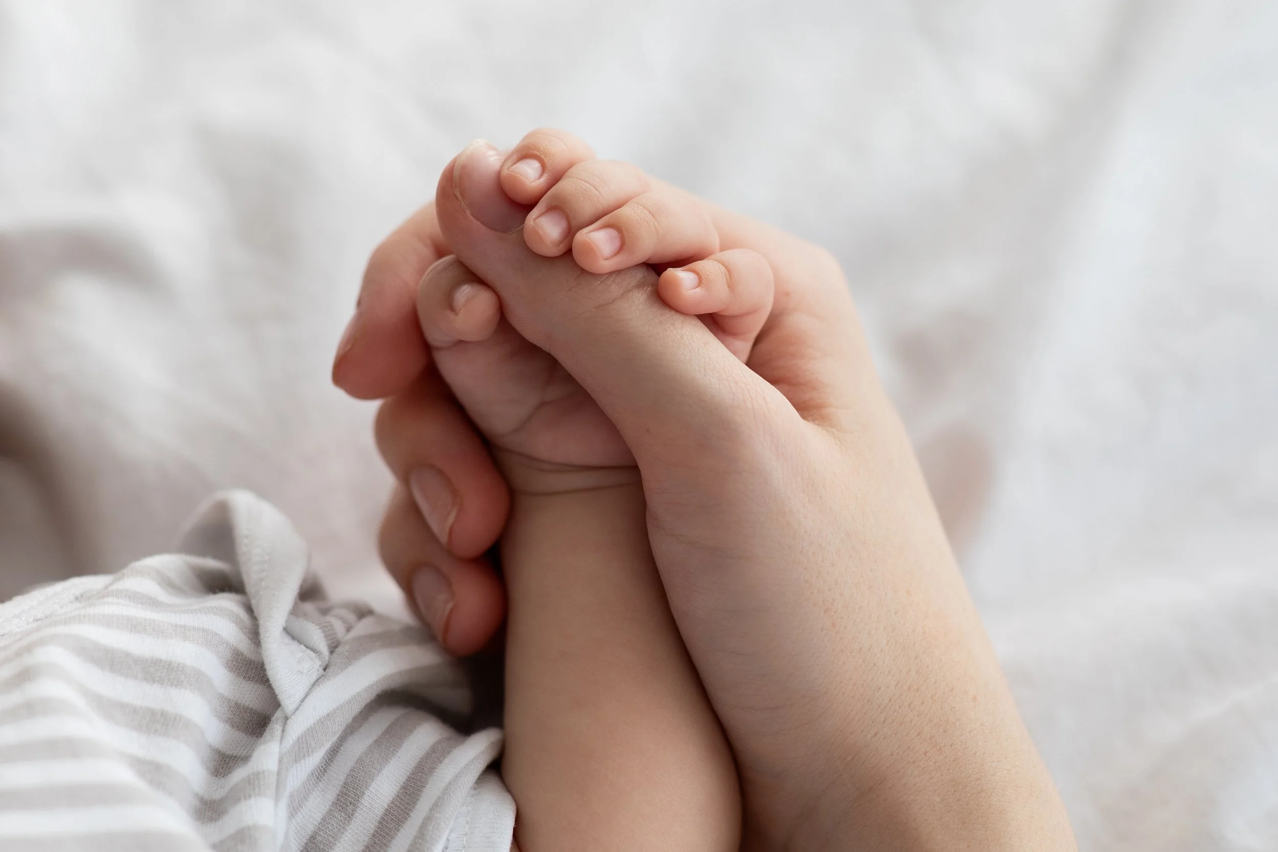 Close-up of an adult and a baby's hands clasped together, with the baby’s hand holding the adult’s finger. The baby is wearing a striped outfit.