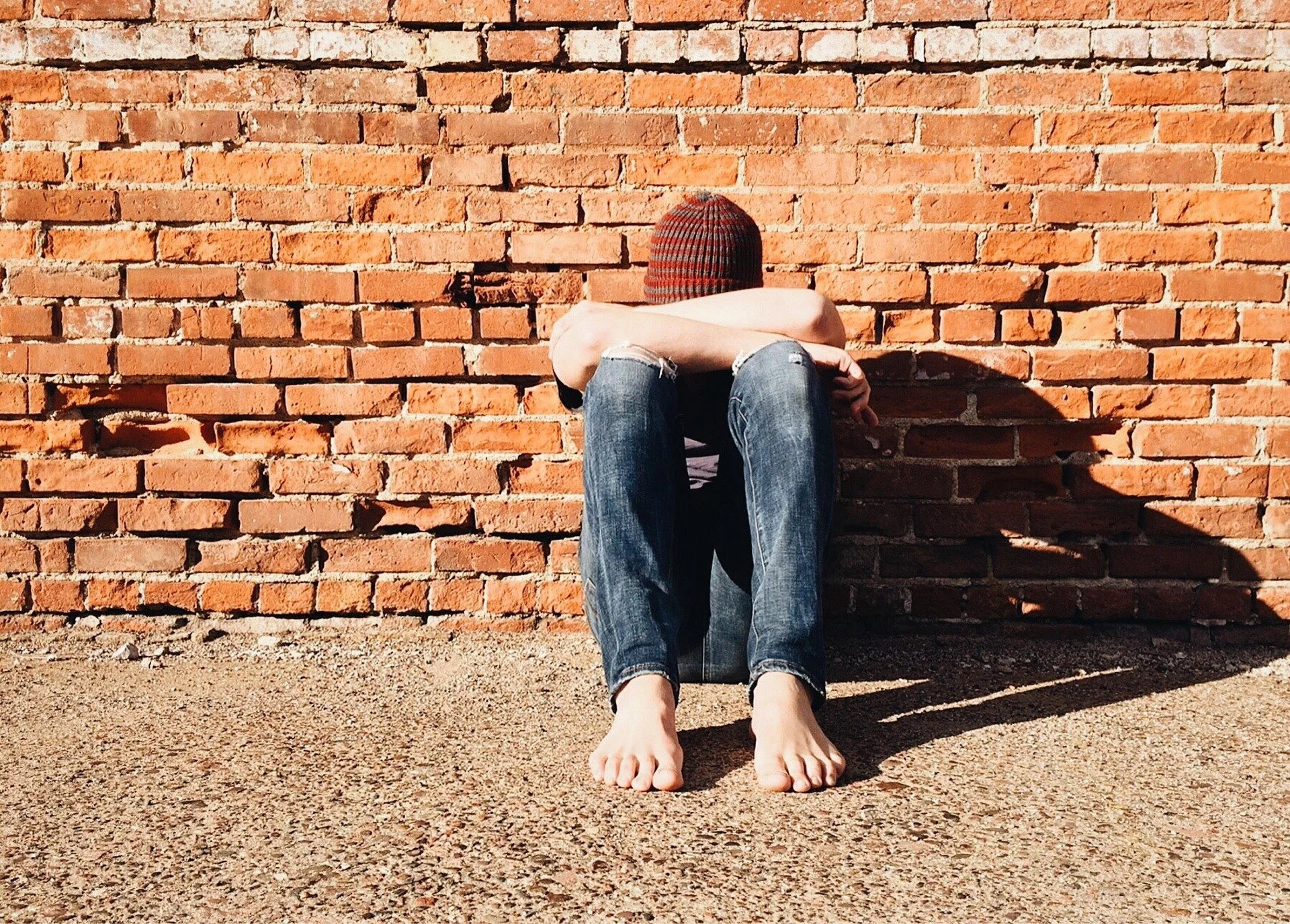 Person sitting against a brick wall, wearing ripped jeans and a beanie, with arms resting on knees and head down.