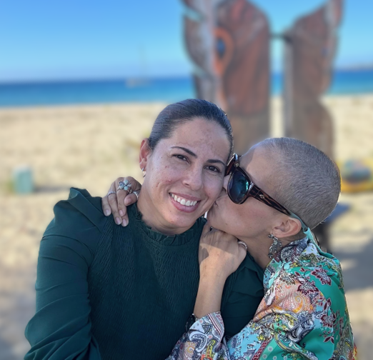 A woman smiling while someone kisses her on the cheek at the beach with a sculpture and ocean in the background.