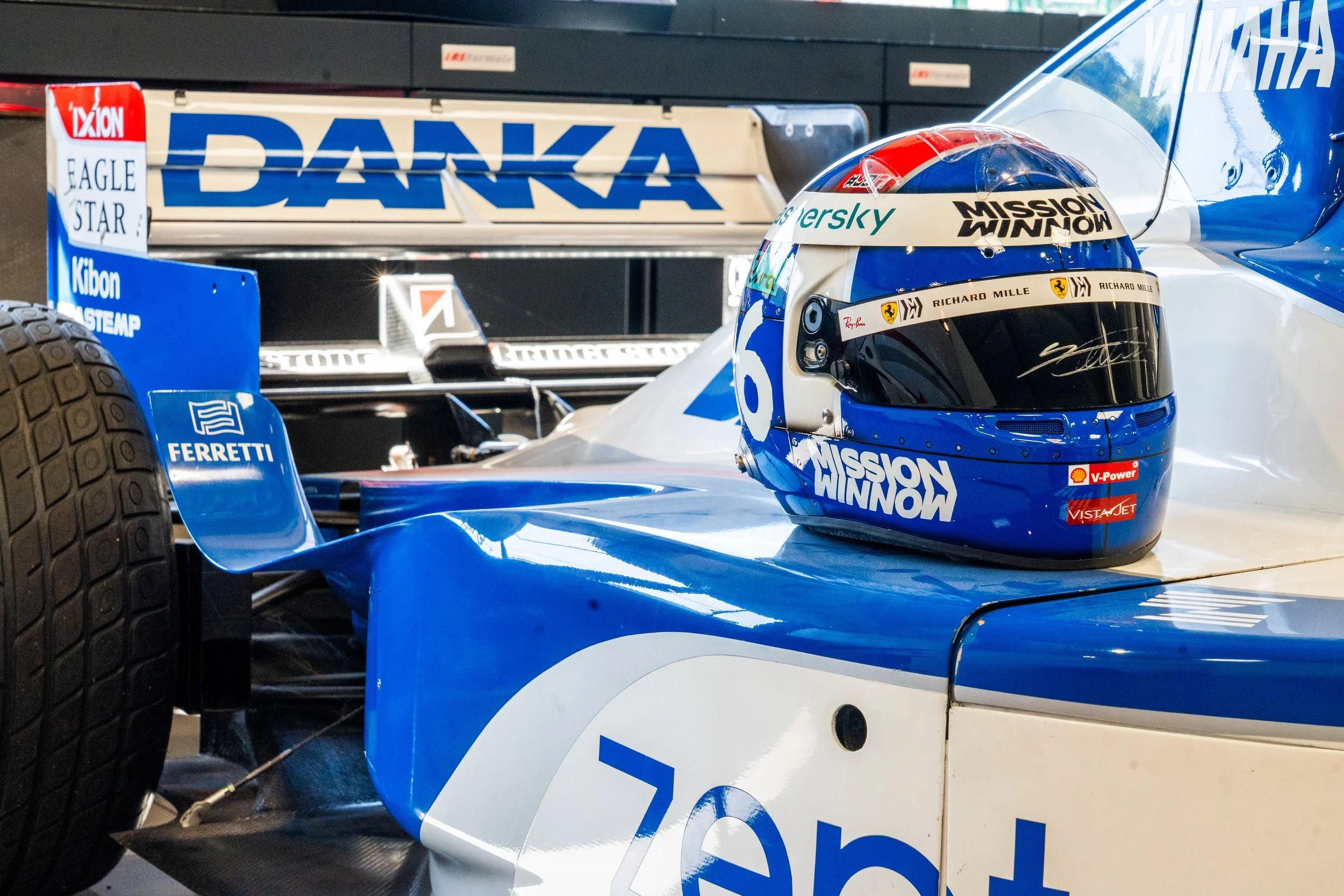Casque de pilote de course de formule 1 placé sur une voiture de course, portant des logos de sponsors et de l'équipe, avec un arrière-plan de partie de la voiture.