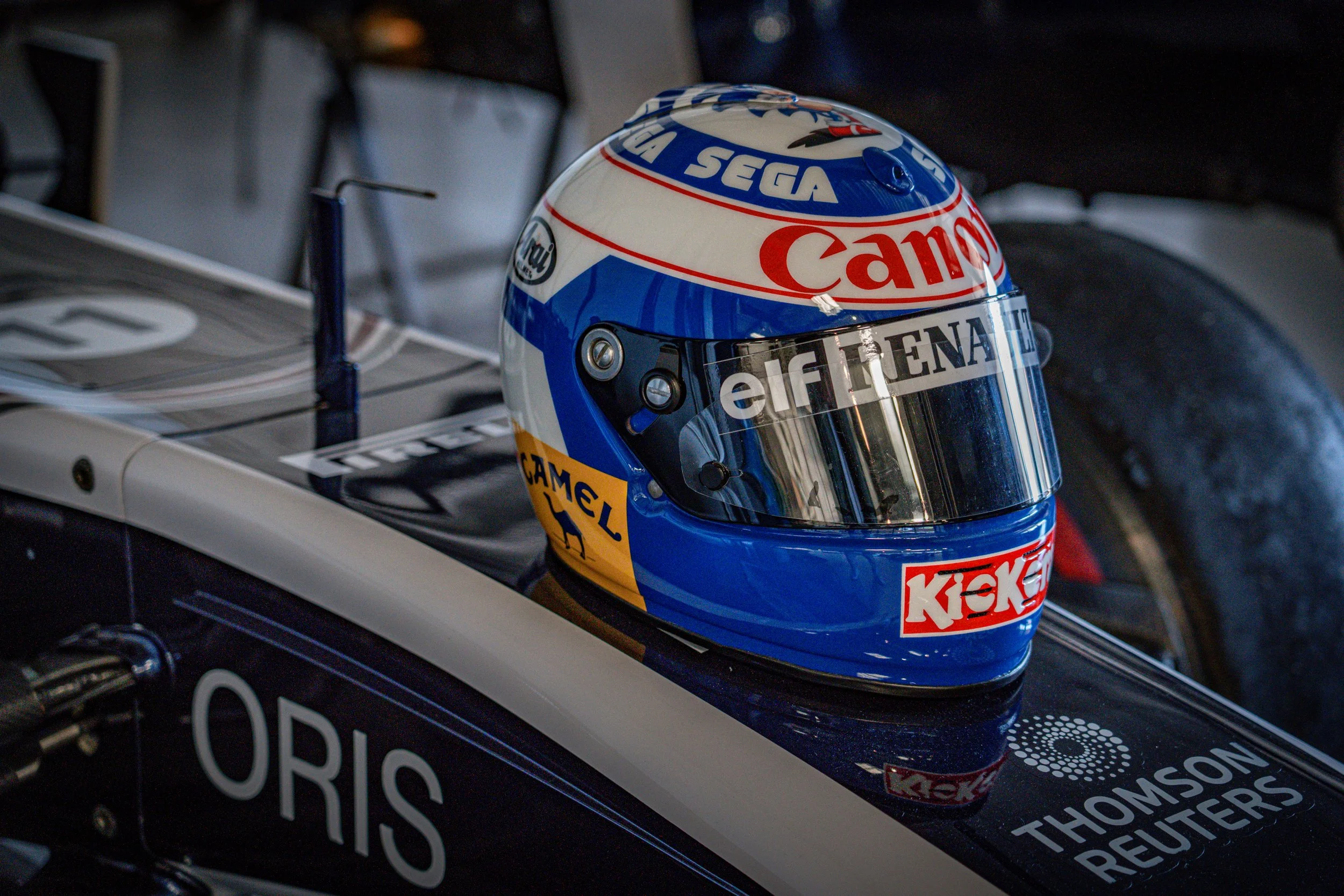 Casque de course de voiture de Formule 1 avec logos de Canon, Elf, Camel, Kickers, et autres, posé sur la voiture.
