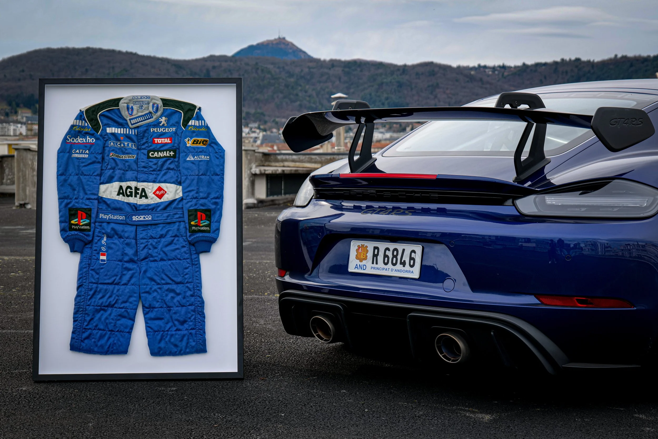 Une voiture de course Porsche 911 GT3 RS noire avec un grand aileron arrière, garée à côté d’un cadre encadrant une combinaison de pilote de course bleue ornée de logos de sponsors. L’arrière-plan montre un paysage urbain avec des montagnes, un ciel