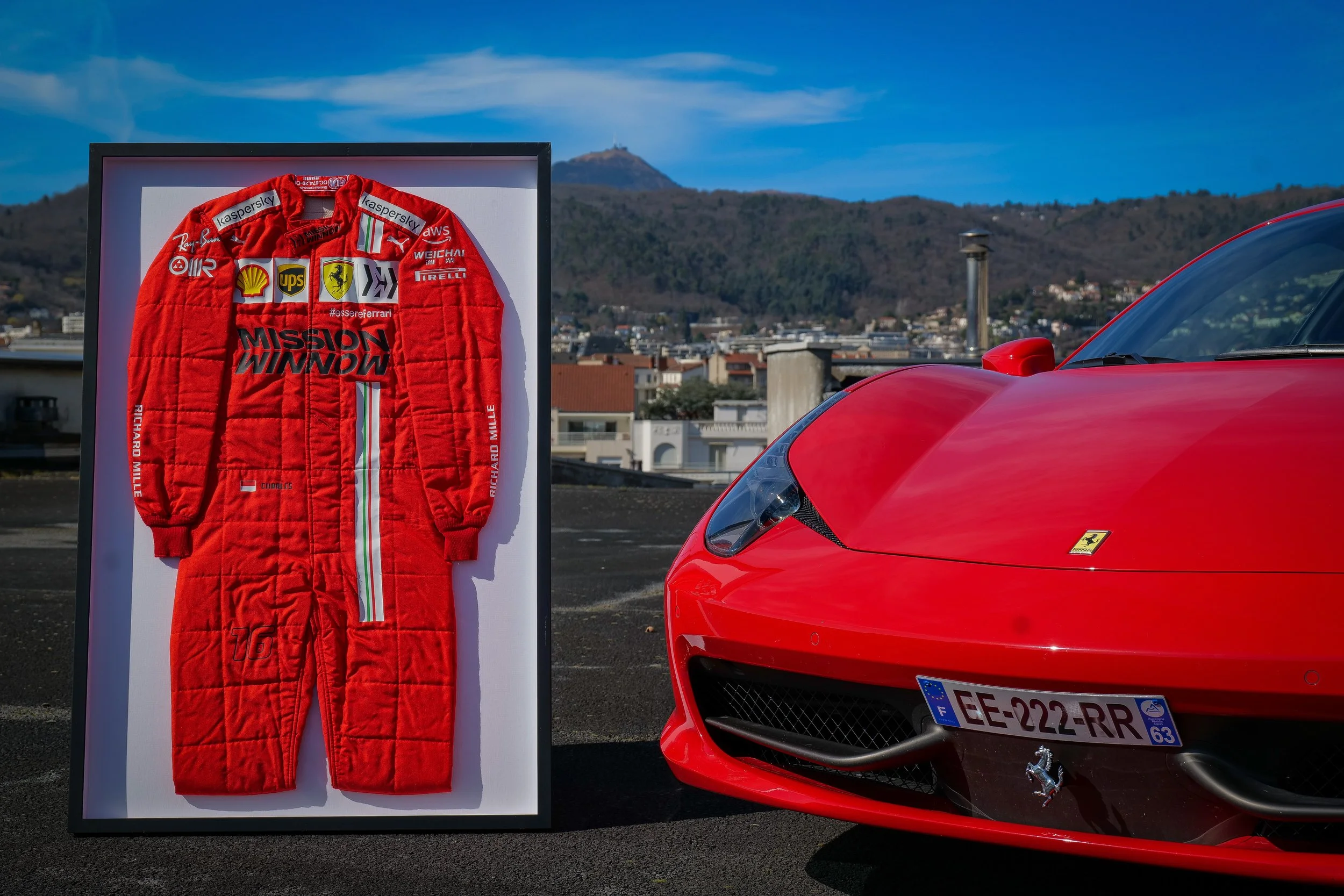 Une combinaison de pilote rouge Ferrari et une voiture Ferrari rouge sur un toit avec une vue urbaine en arrière-plan.