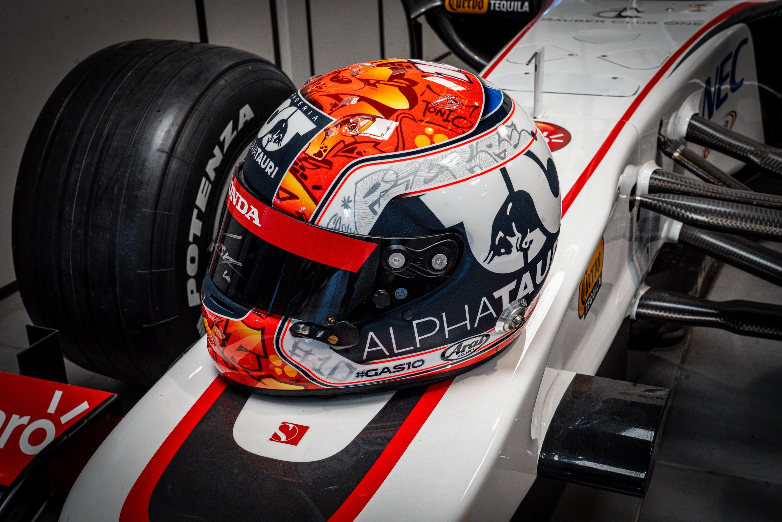Casque de course de formule 1 blanc avec des détails rouges, noirs et orange, portant le logo ALPHATAURI, placé sur une voiture de course de formule 1 blanche avec des toiles d'araignée stylisées, dans un garage.