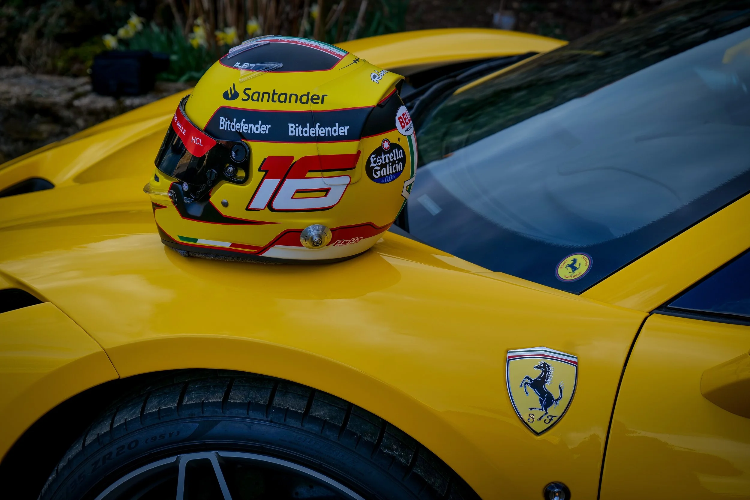 Casque de course Ferrari jaune avec le numéro 16, posé sur la capot d'une voiture Ferrari jaune, avec le logo Ferrari visible sur le côté.