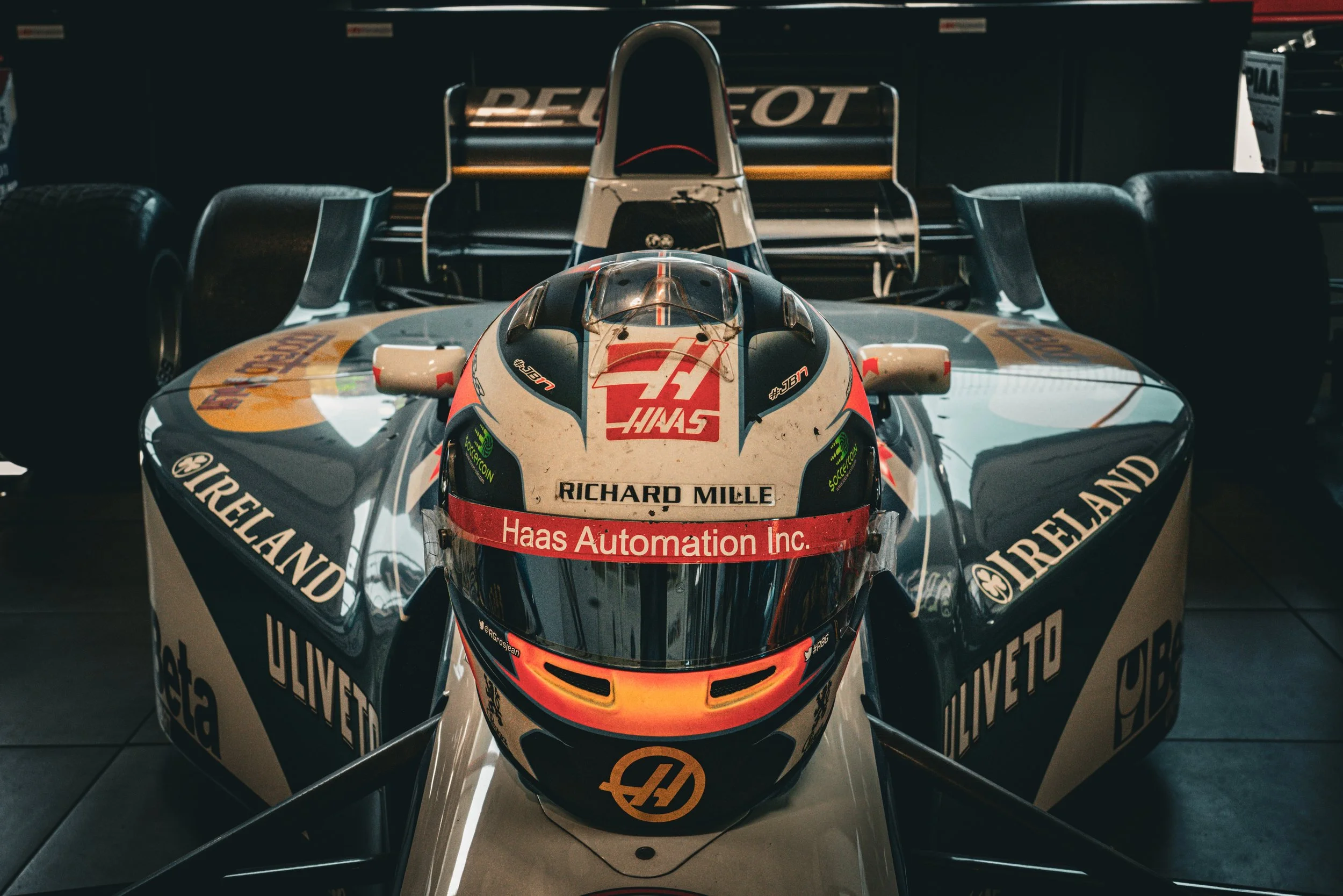 Casque de course de voiture de Formule 1 avec logos de Haas, Richard Mille, Aero, et Ireland, placé sur une voiture de course dans un garage ou showroom.