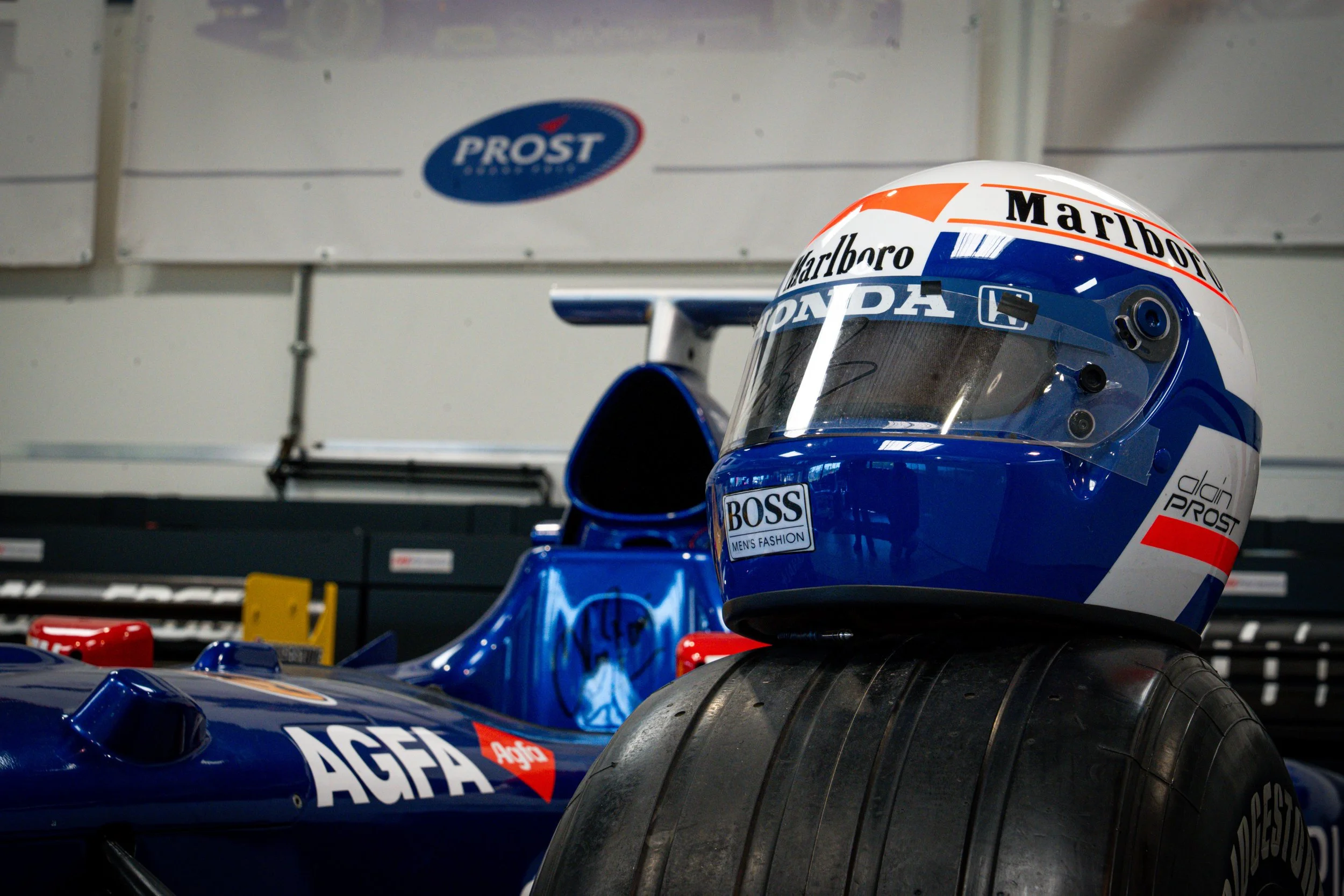Un casque de pilote de F1 posé sur une roue avant d'une voiture de course de Formule 1 dans un garage, avec des logos de sponsors et une voiture de course bleue.