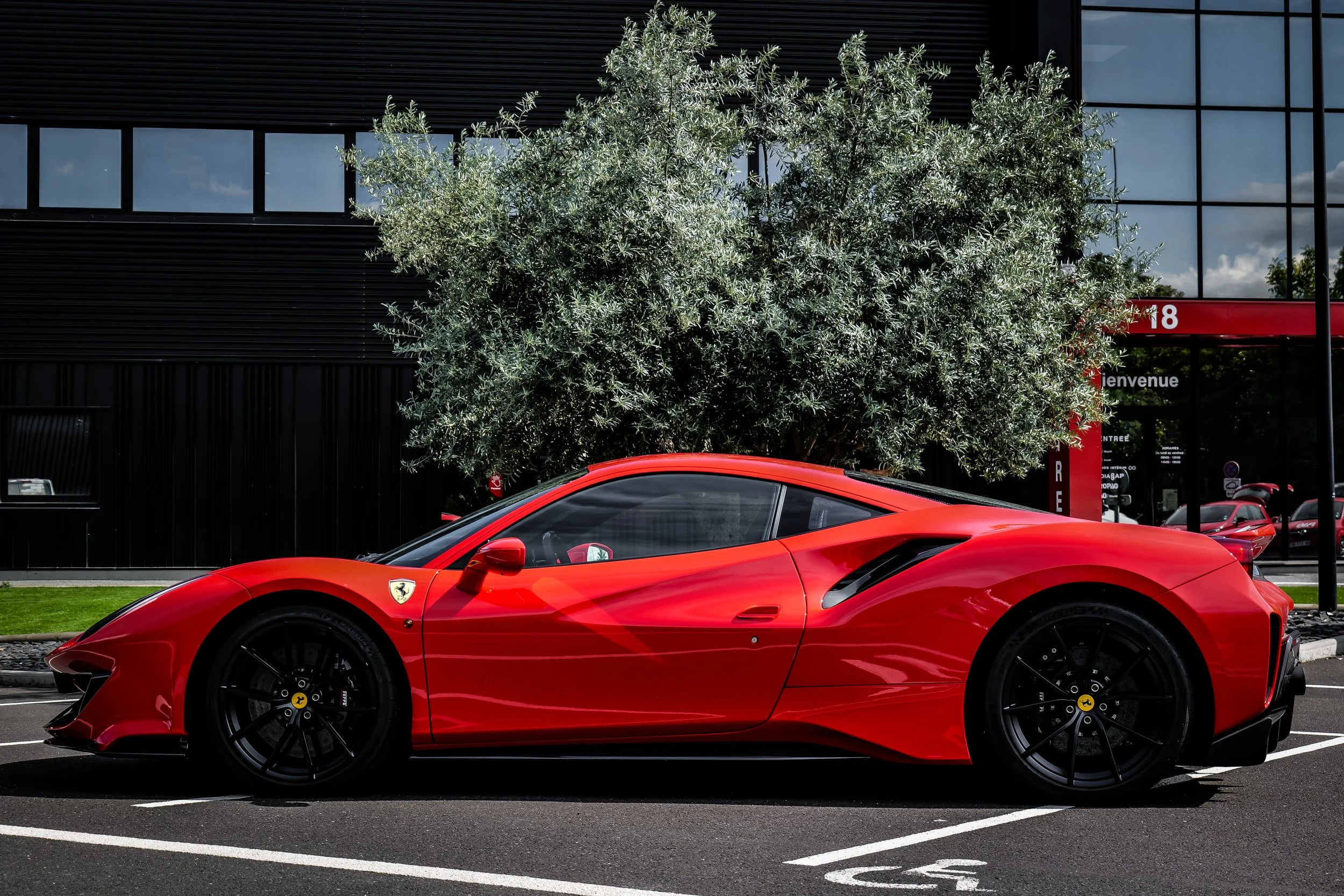Une voiture de sport Ferrari rouge garée dans un parking devant un bâtiment moderne avec des fenêtres en verre et une grande plante verte en arrière-plan.