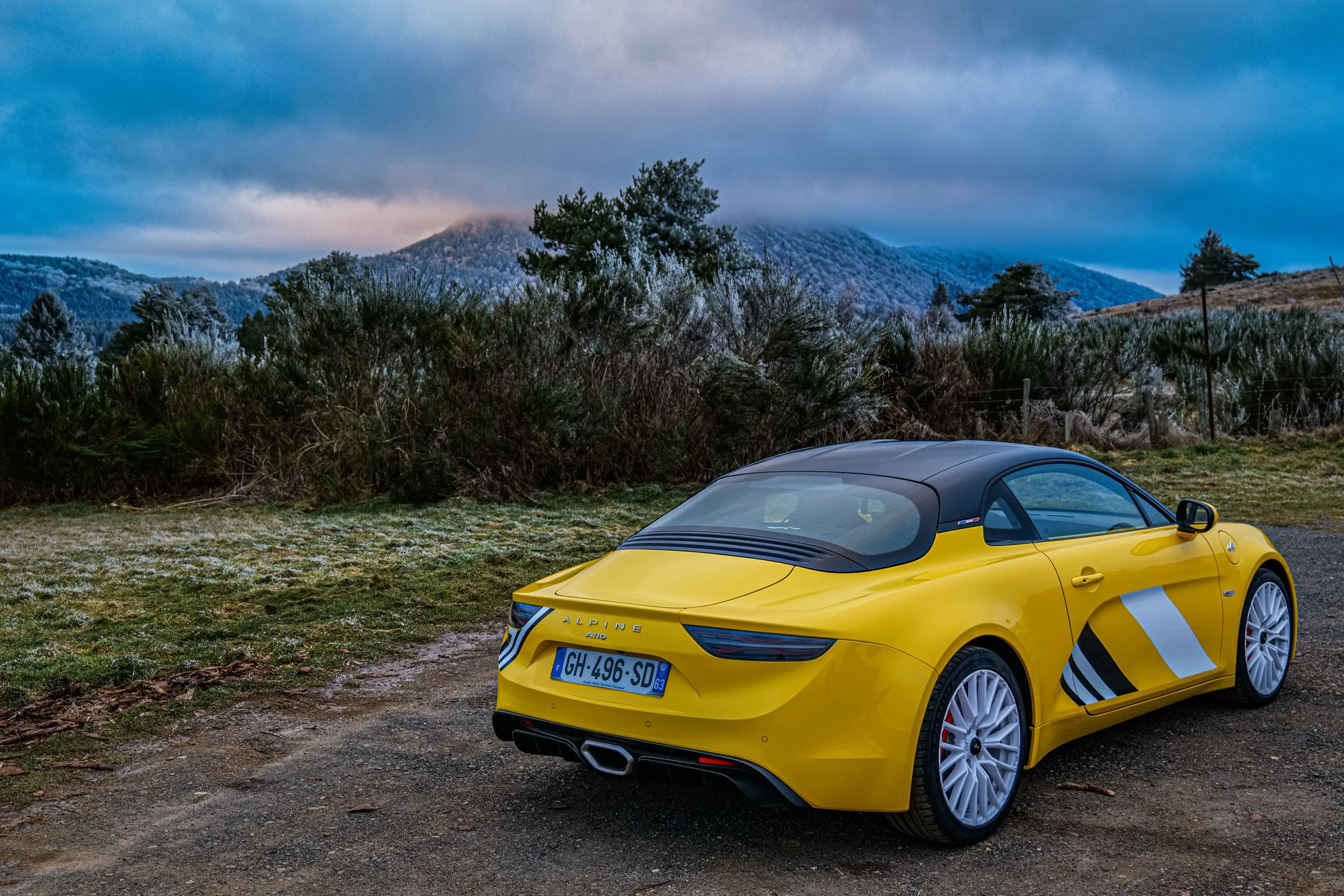 Voiture jaune décapotable Alpine avec toit noir, stationnée sur un chemin peu pavé, arrière-plan montagneux et végétation sous un ciel nuageux.