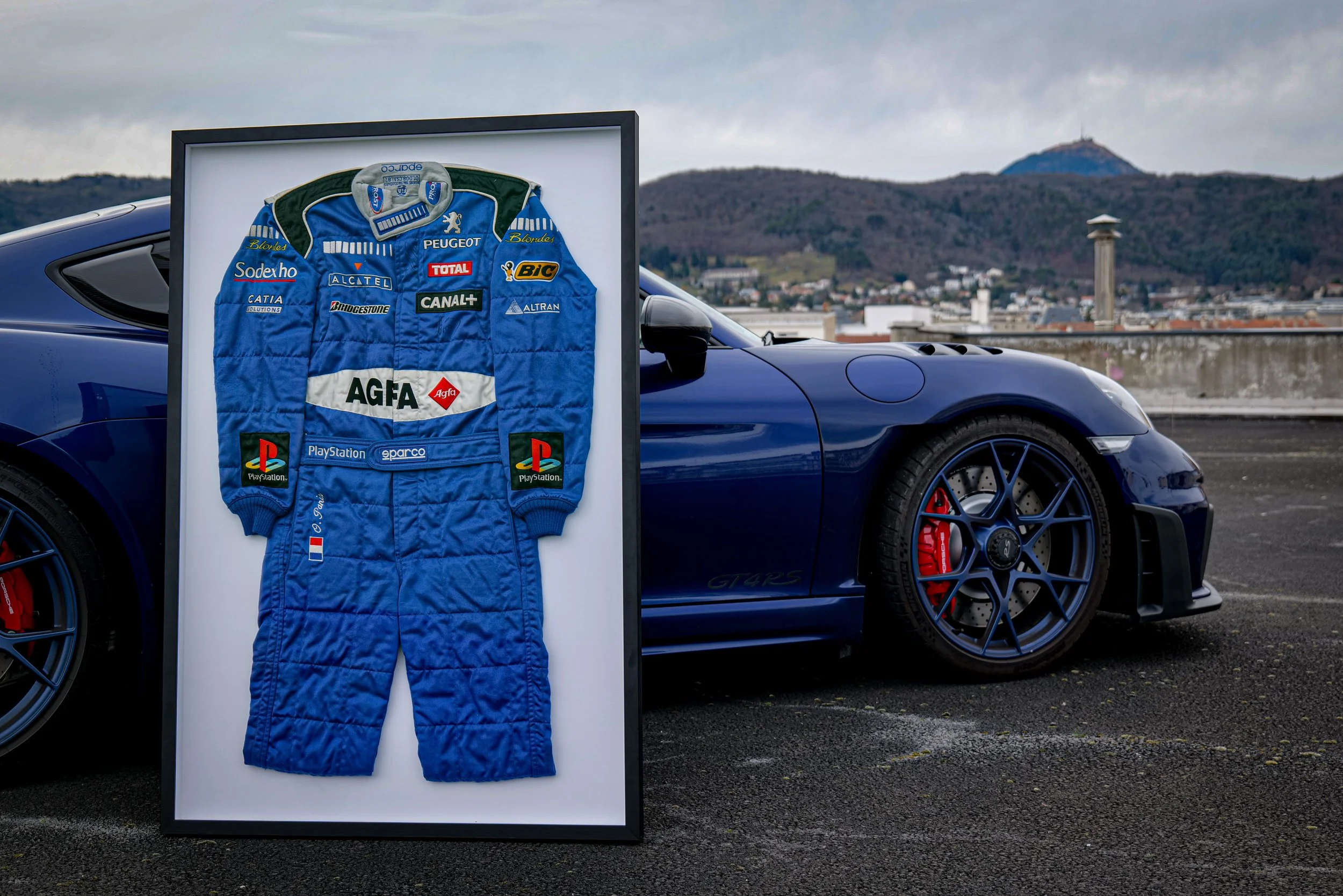 Une combinaison de course bleue avec des logos de sponsors, encadrée, posée contre une voiture de sport bleue sur un parking avec un paysage urbain en arrière-plan.