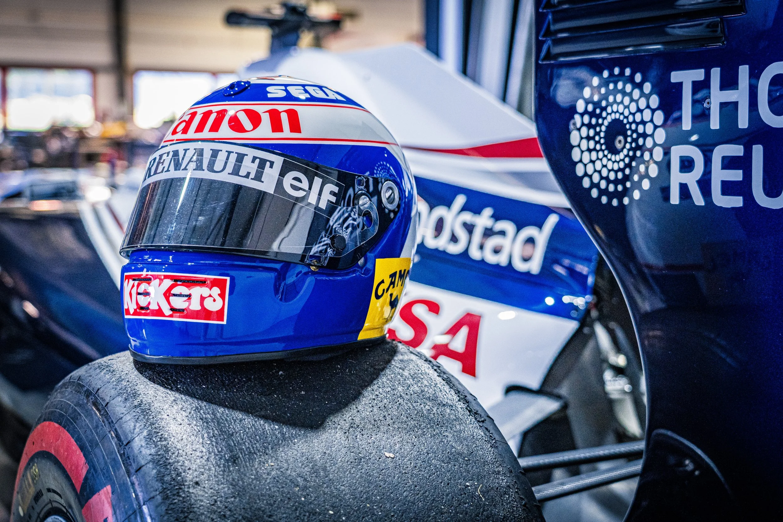 Casque de course de Formule 1 avec logos sponsor, posé sur une roue de voiture de course dans un garage.