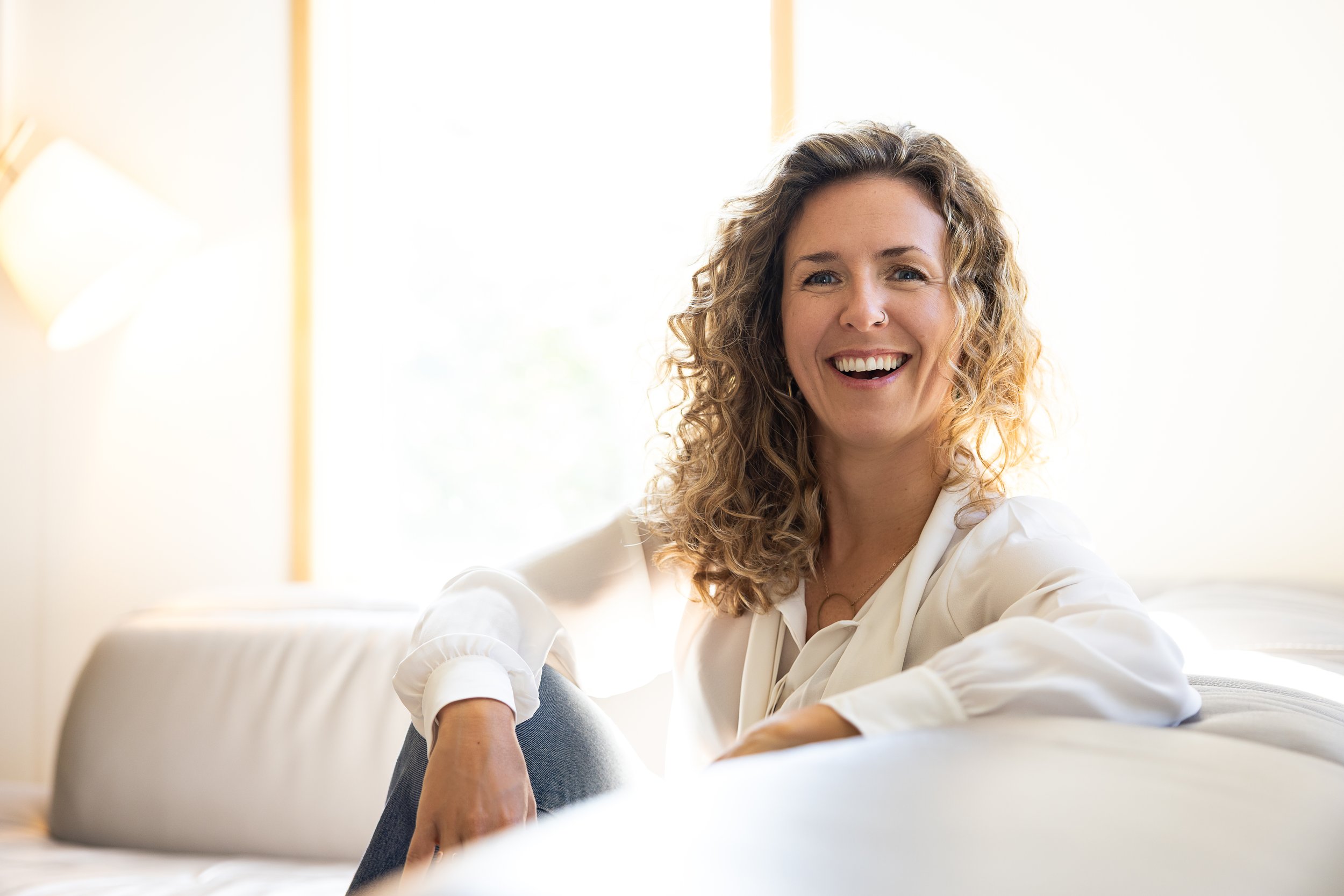 Une femme souriante avec des cheveux bouclés, portant une chemise blanche, assise sur un canapé dans une pièce lumineuse.