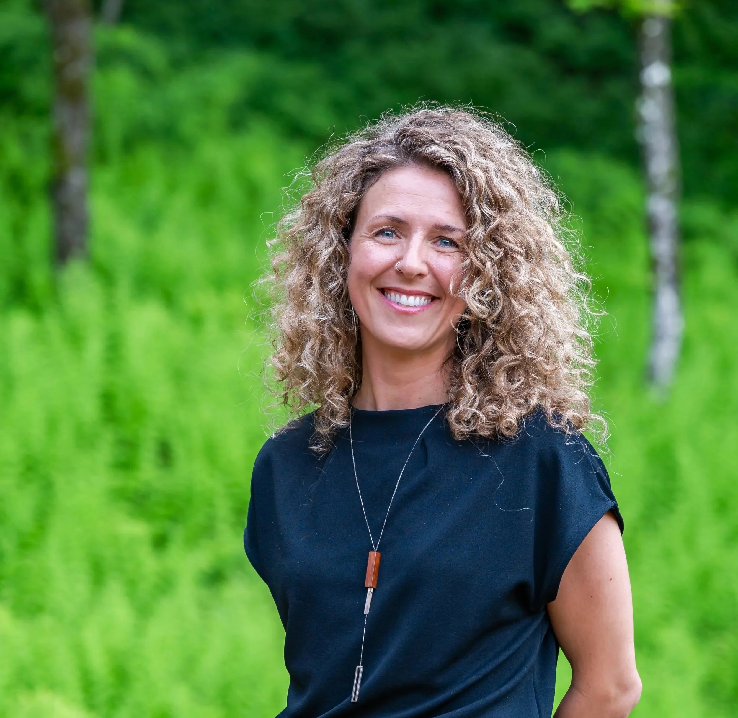 Une femme souriante avec des cheveux bouclés, portant un t-shirt noir et un collier, se tient devant un fond de nature vert avec des arbres.