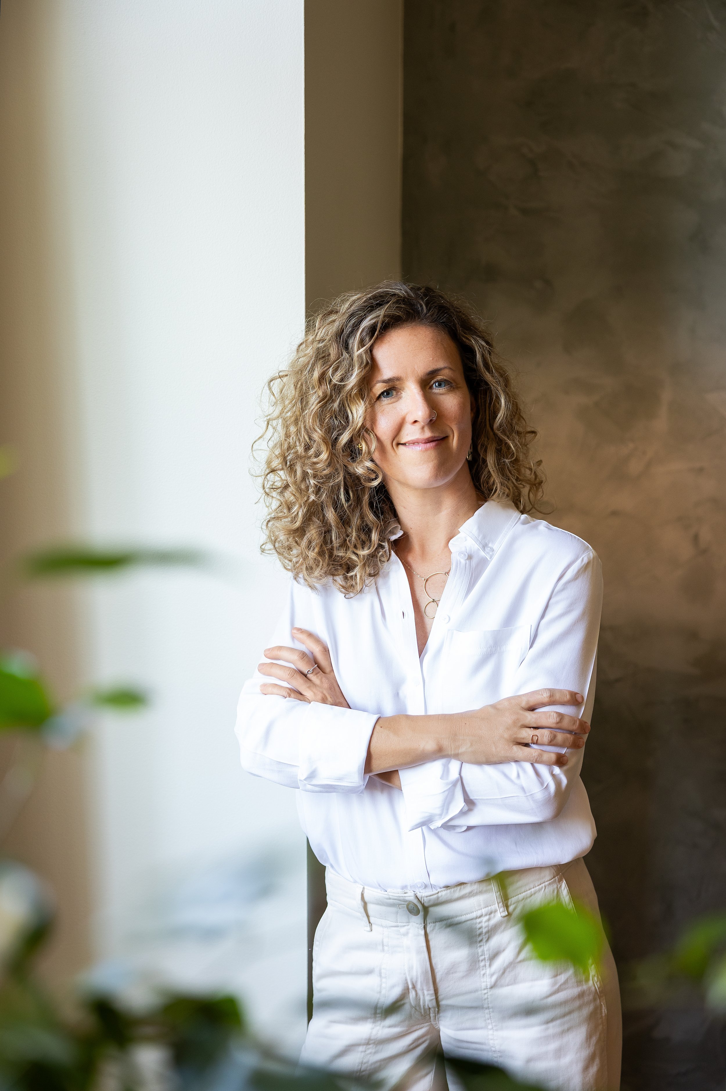Femme aux cheveux bouclés, portant une chemise blanche, debout près d'une fenêtre avec un mur marron en arrière-plan