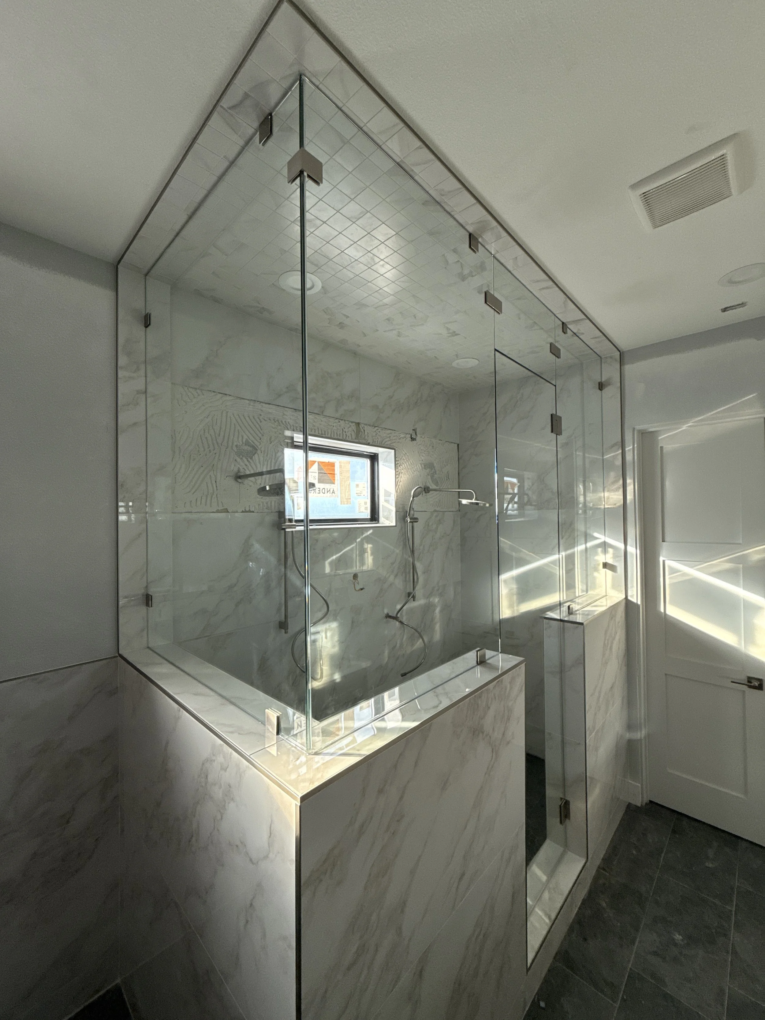 A glass shower enclosure with brass hardware in a bathroom under construction, with cabinetry on the left and a wall with markings on the right.